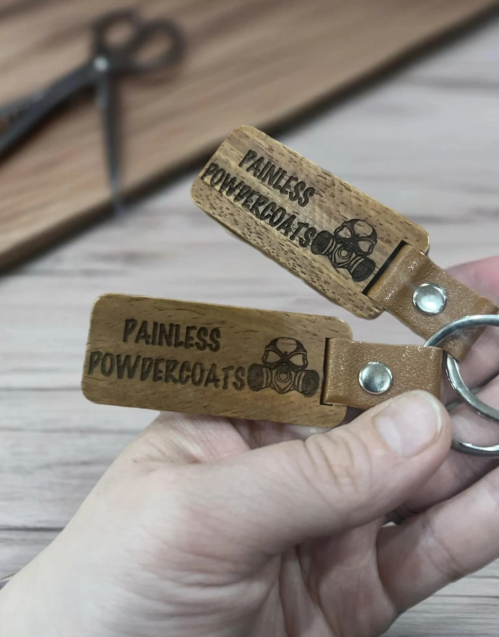 Person holding custom wooden keychains with Painless Powdercoats logo and text.