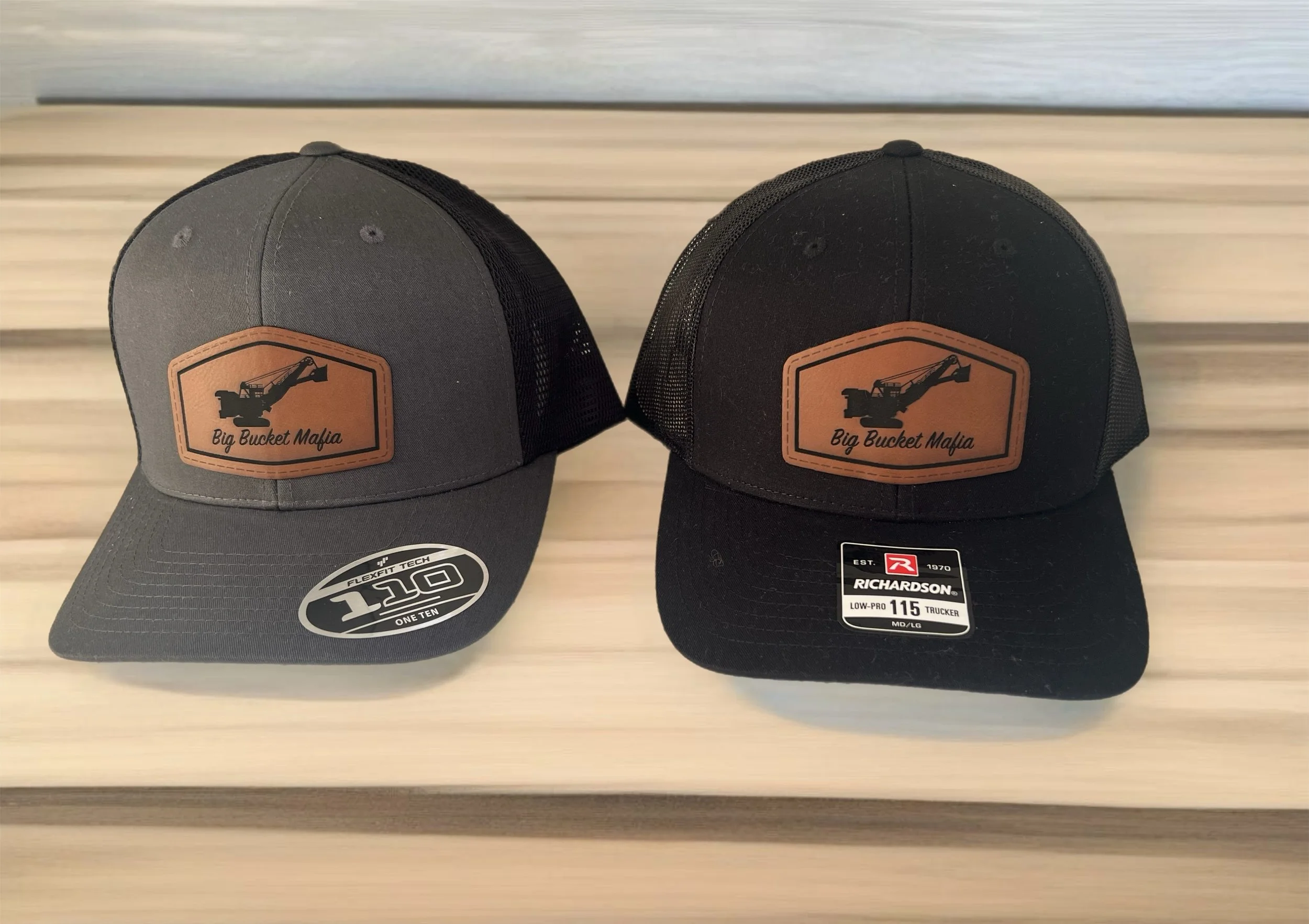 Two caps with mesh back, one gray and one black, featuring a "Big Bucket Mafia" logo with an excavator silhouette, on a wooden surface.