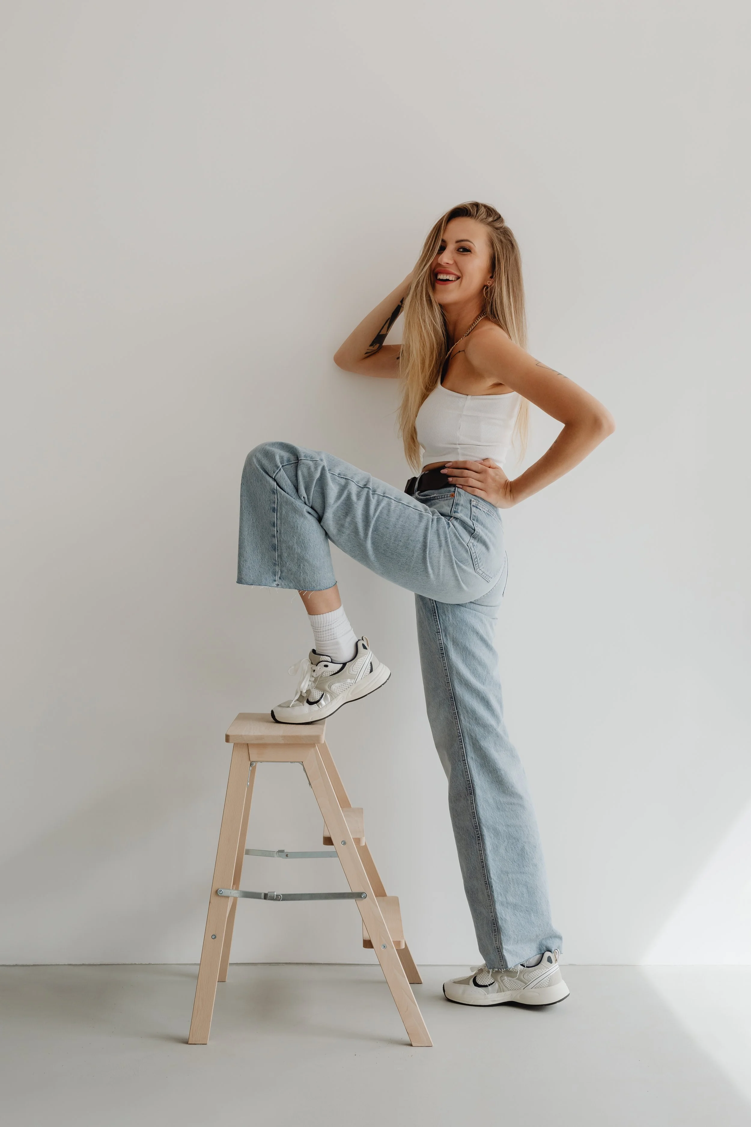 A young woman with long blonde hair, wearing a white sleeveless top, light blue jeans, and white sneakers, standing with one leg on a step ladder, smiling and posing against a plain white wall.