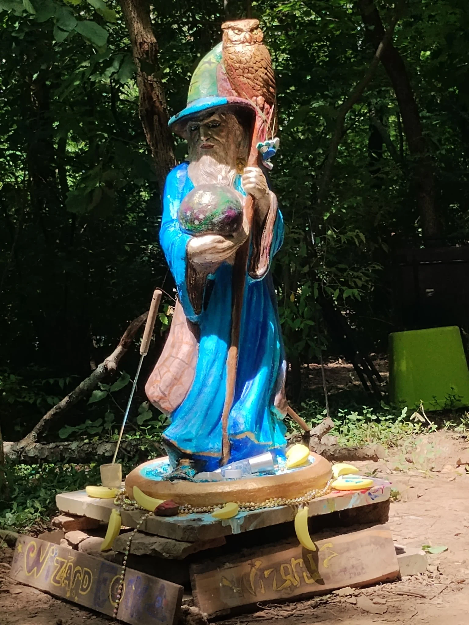 Colorful wooden sculpture of a wizard holding a staff, with a large fish in one hand, wearing a blue robe and a tall hat, decorated with various objects including a bird and symbolic ornaments, set outdoors among trees.