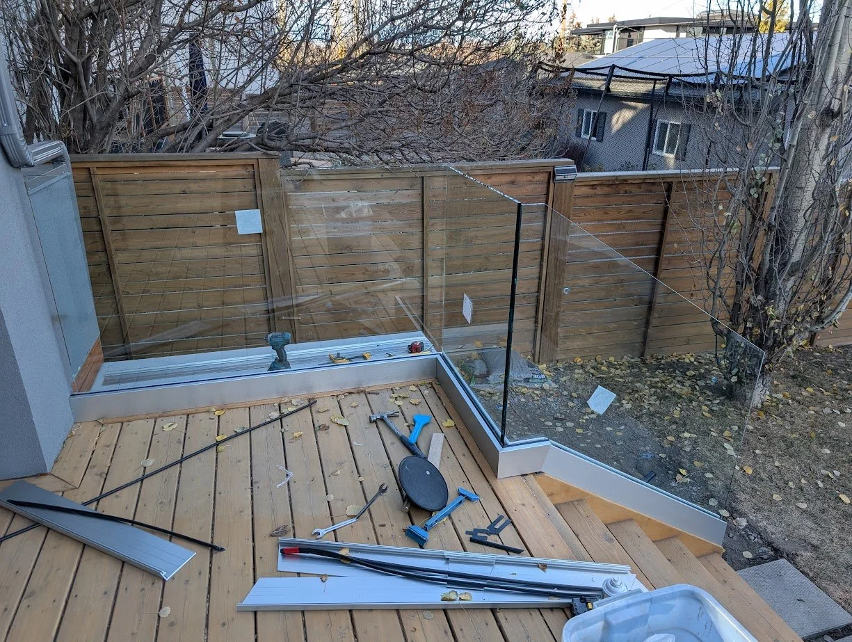 A partially assembled glass railing on a wooden deck with construction tools, including a drill, hammer, and metal strips, in a backyard enclosed by a wooden fence, with trees and neighboring houses visible in the background.