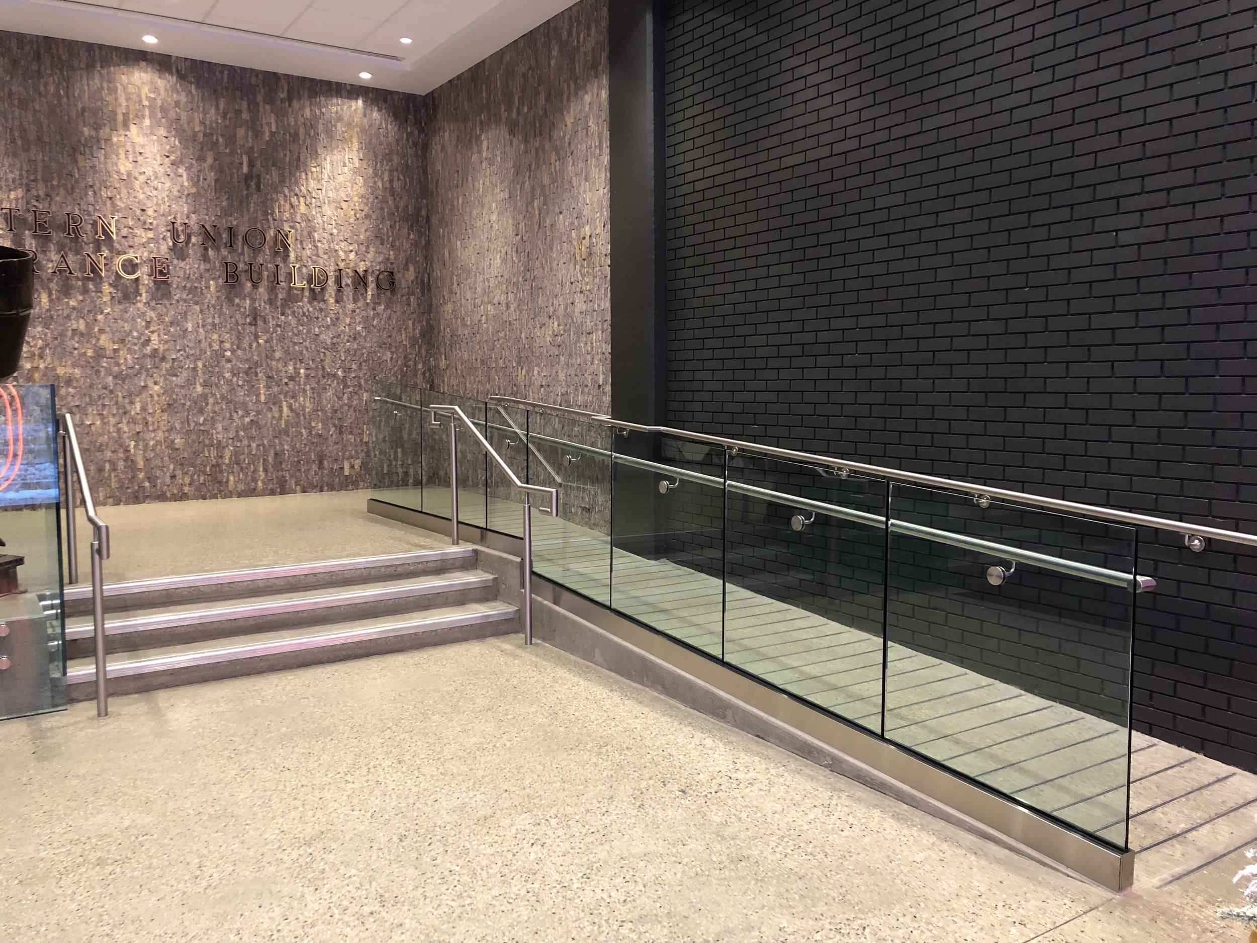 Interior of a building entrance with stairs and a wheelchair ramp, black brick wall, glass railings, and the sign 'Eastern Union Finance Building' on the wall.