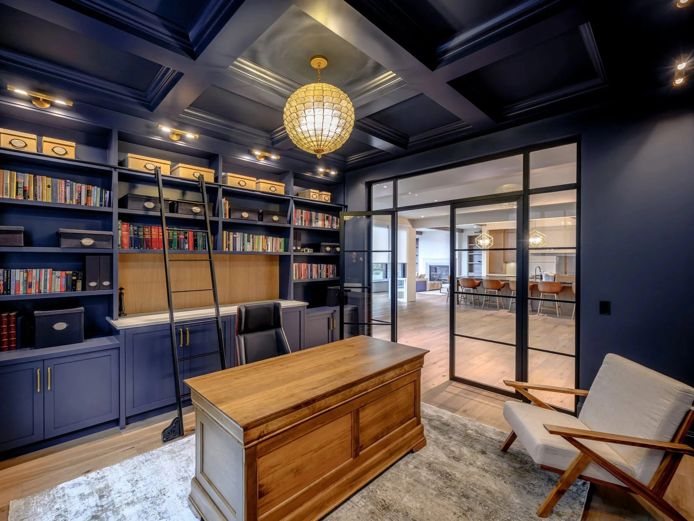 Modern home office with blue built-in bookshelves, a wooden desk, a black office chair, a white armchair, and glass-paneled doors leading to a room with a kitchen counter and bar stools.