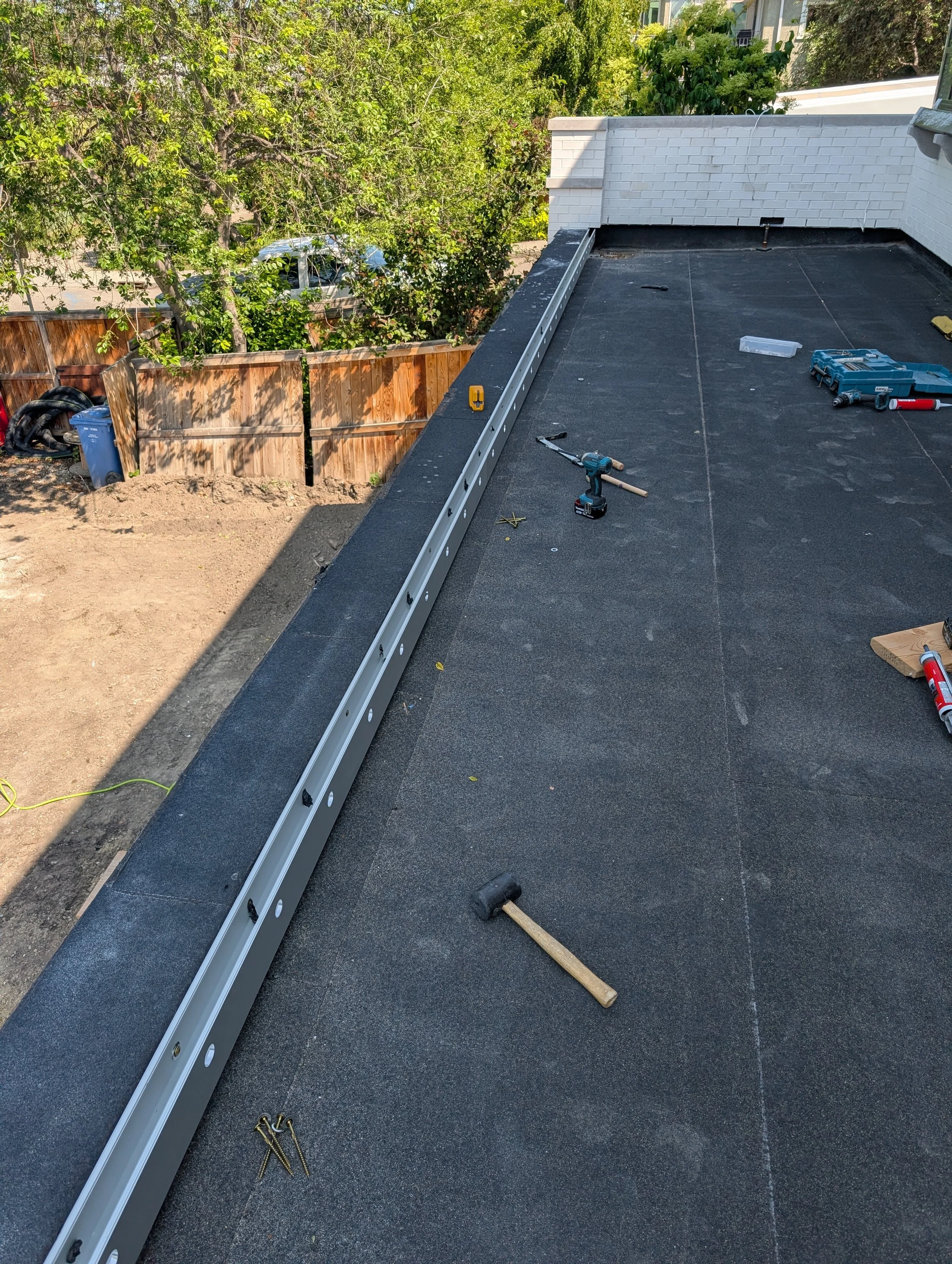 A rooftop under construction with tools and materials, a metal beam installed along the edge, and a black surface. There are trees, a wooden fence, and parts of a house visible in the background.