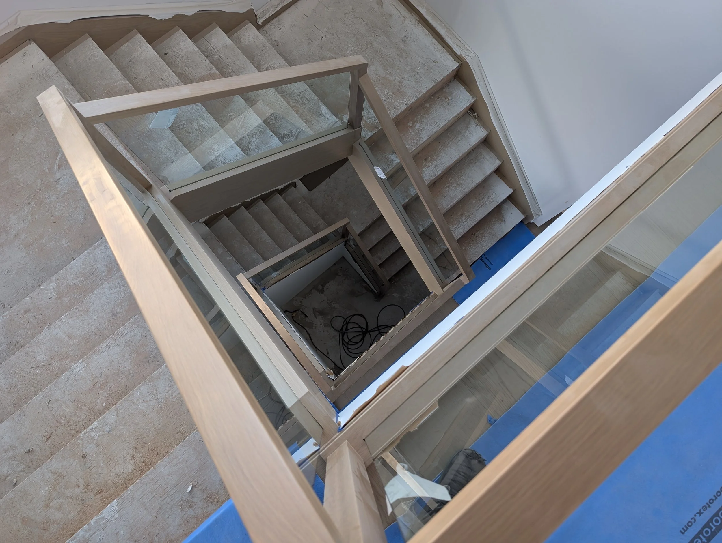 View looking down through a glass railing onto a staircase with wooden handrails and beige steps, showing multiple levels and electrical cords on the ground at the bottom.