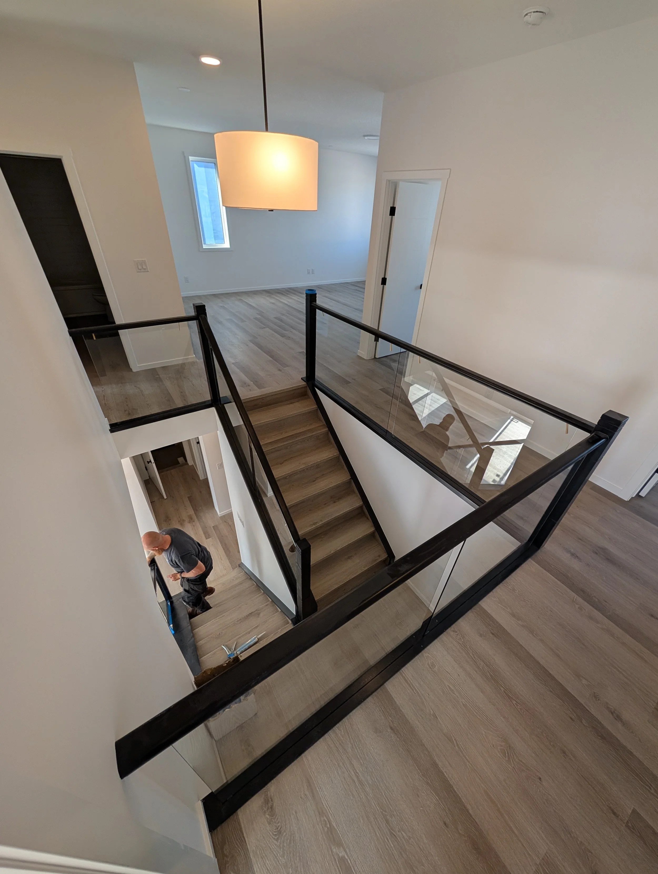 View of an interior staircase with wooden steps and glass railing, overlooking the lower level of a modern home with light-colored walls, wooden flooring, and a large pendant light hanging from the ceiling.