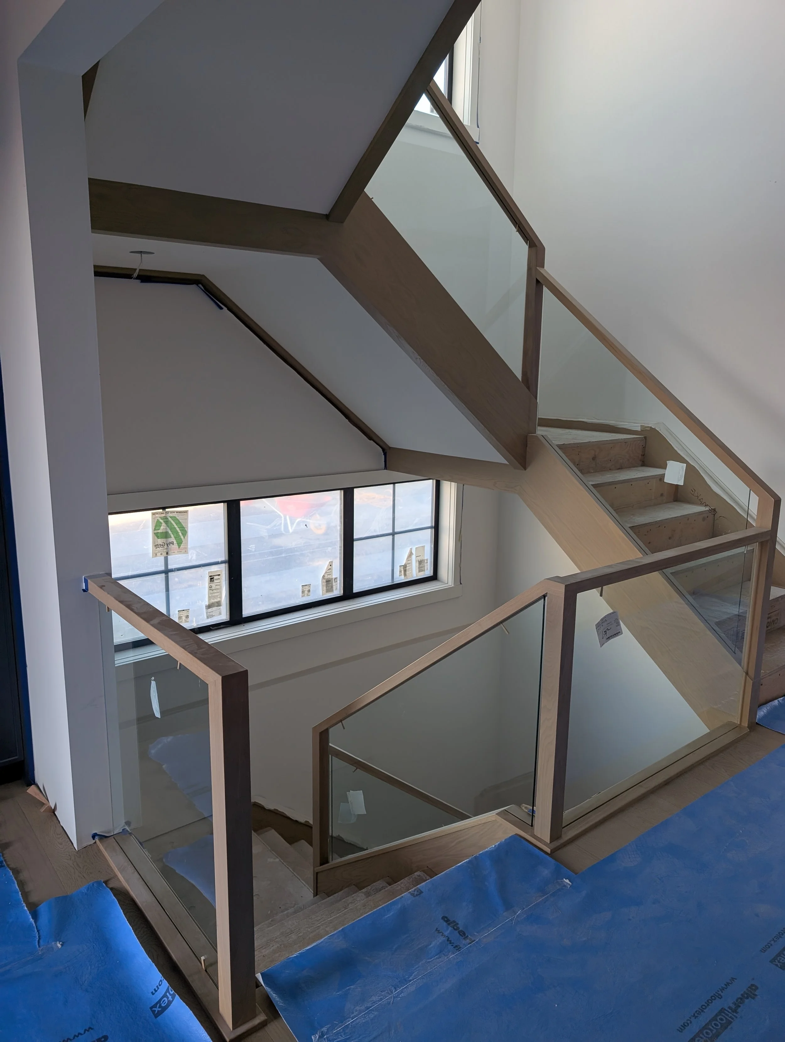 Interior view of a staircase with wooden railings and glass panels, under construction, with a window in the background and blue protective covering on the floor.
