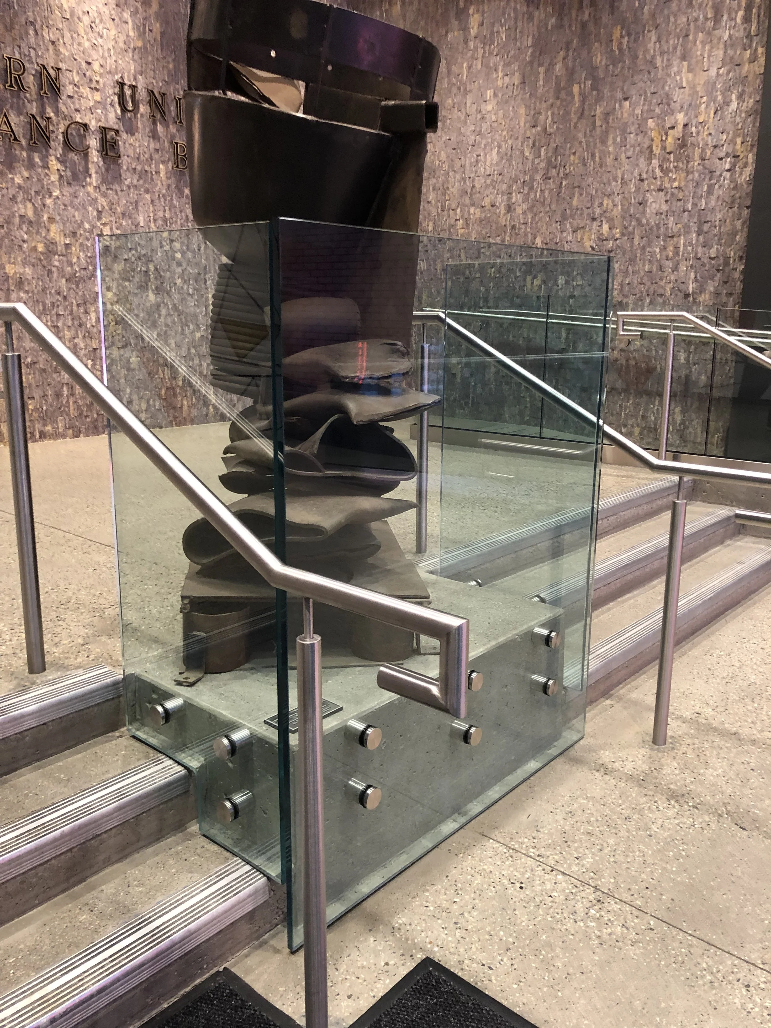 Decorative metal sculpture behind a glass barrier in a public building, with stairs and a textured wall in the background.