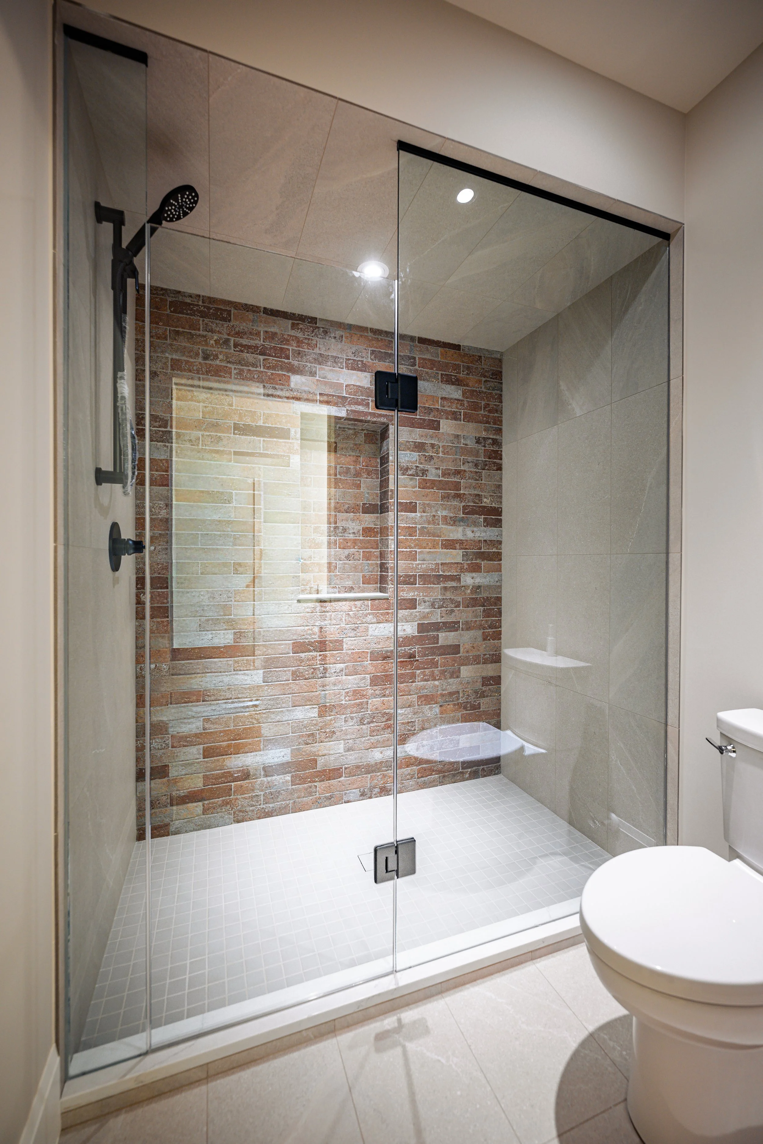 Modern bathroom with a glass-enclosed shower featuring a brick wall inside and beige tiles on the floor, next to a white toilet.
