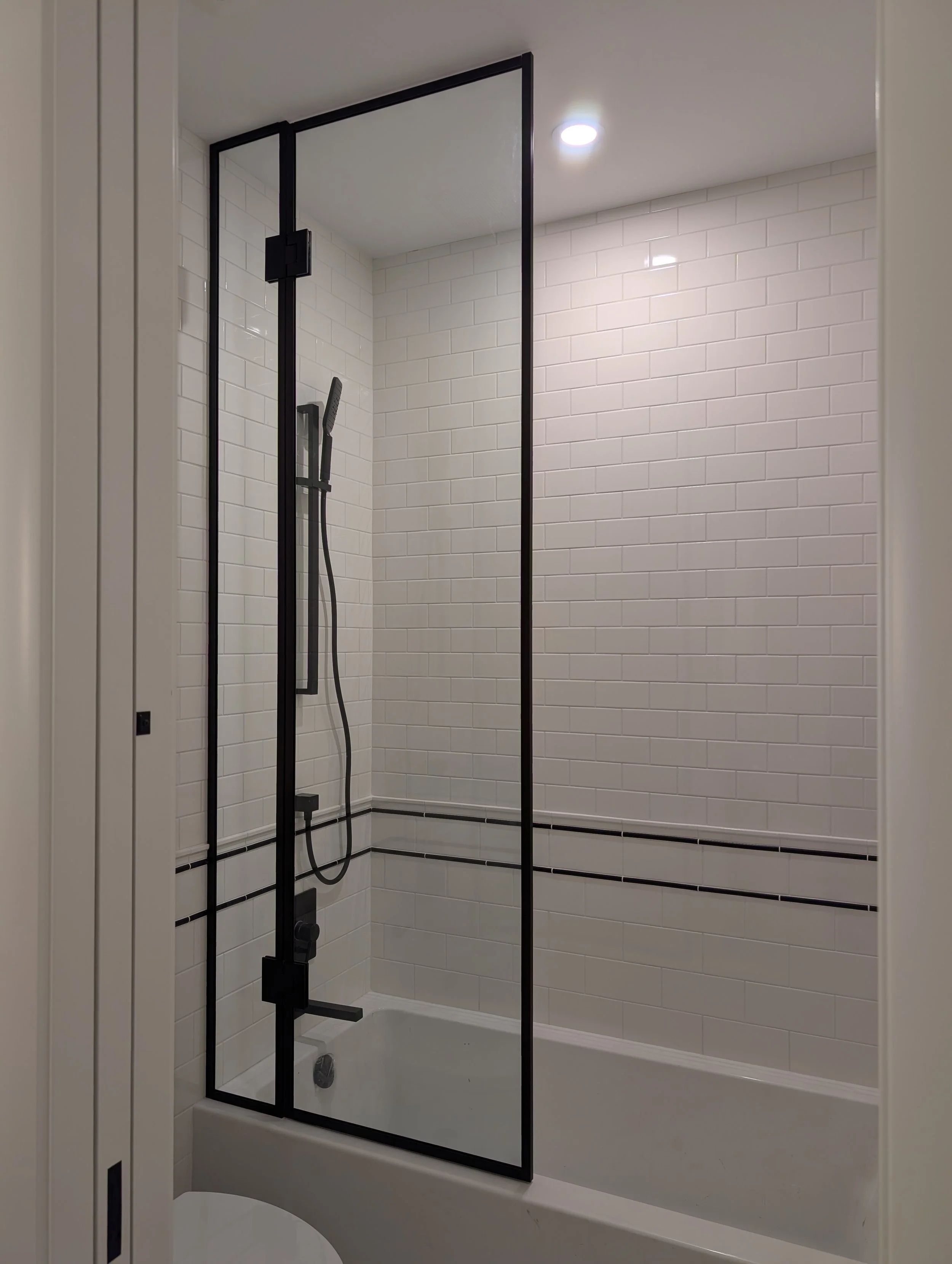 Modern bathroom with a walk-in shower enclosed by black-framed glass, white subway tile walls, and a ceiling-mounted light.
