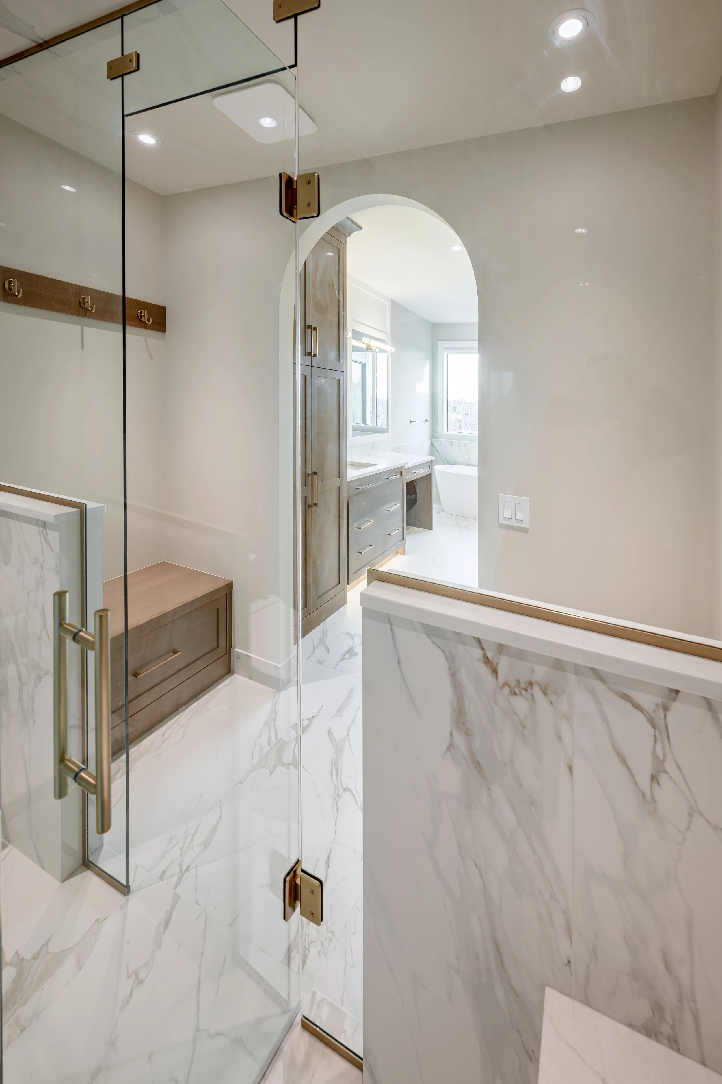 Modern bathroom with glass shower enclosure, marble flooring, wooden accents, and a view of a bathtub through an arched doorway.