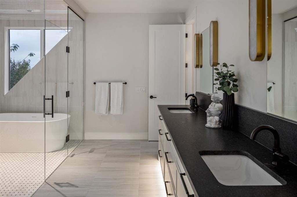 A modern bathroom with a black countertop vanity holding a vase with greenery and decorative jars. Beside the vanity is a mirror with gold accents. There is a white door and two white towels hanging on the wall. In the background, a bathtub is enclos