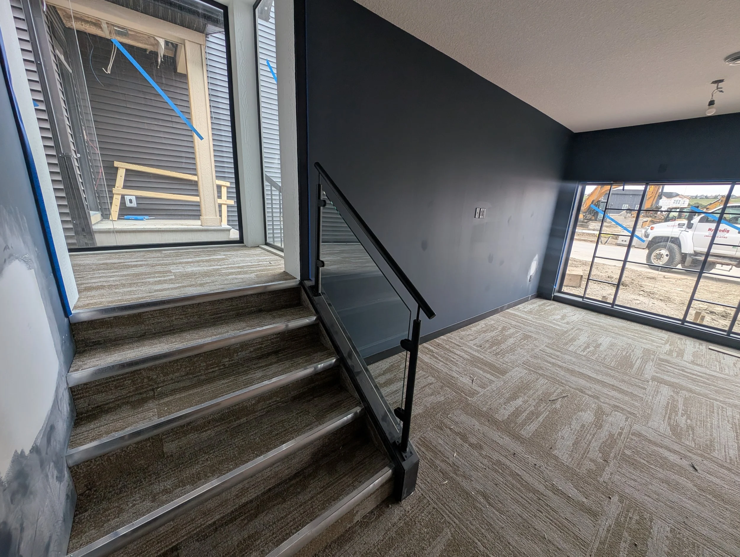 Interior of a building under construction with new carpet and stairs leading to a second floor, large windows showing construction equipment outside, and a black wall.