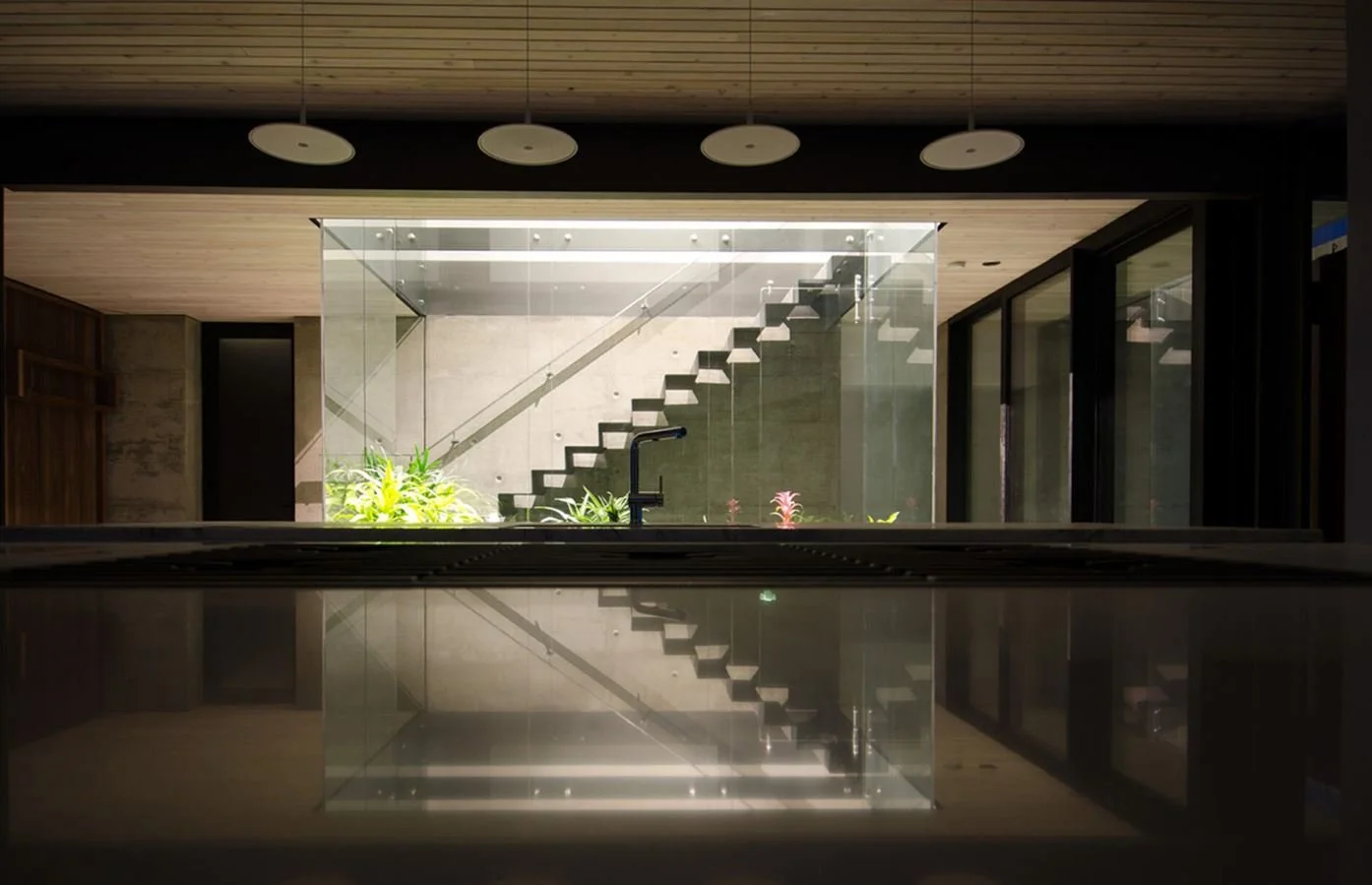 Interior view of a modern home with a staircase seen through a large glass wall, reflected on a glossy surface below, with a black faucet and green plants near the window.