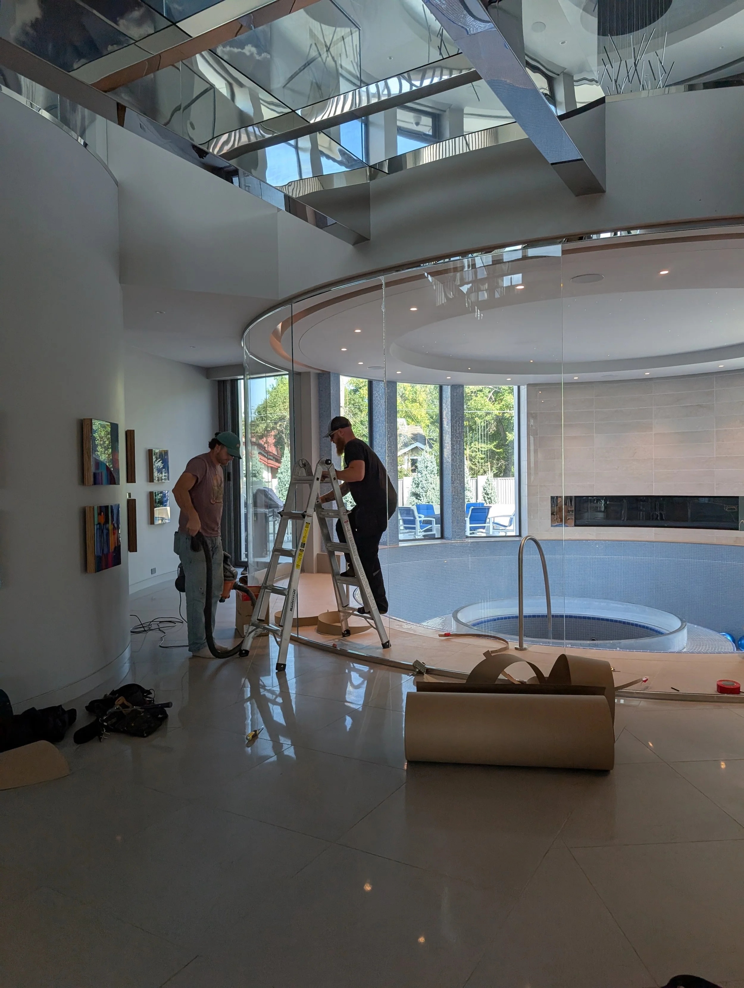 Two workers preparing an indoor pool area, with one standing on a ladder and the other next to a vacuum cleaner, inside a modern house with large windows and artwork on the wall.