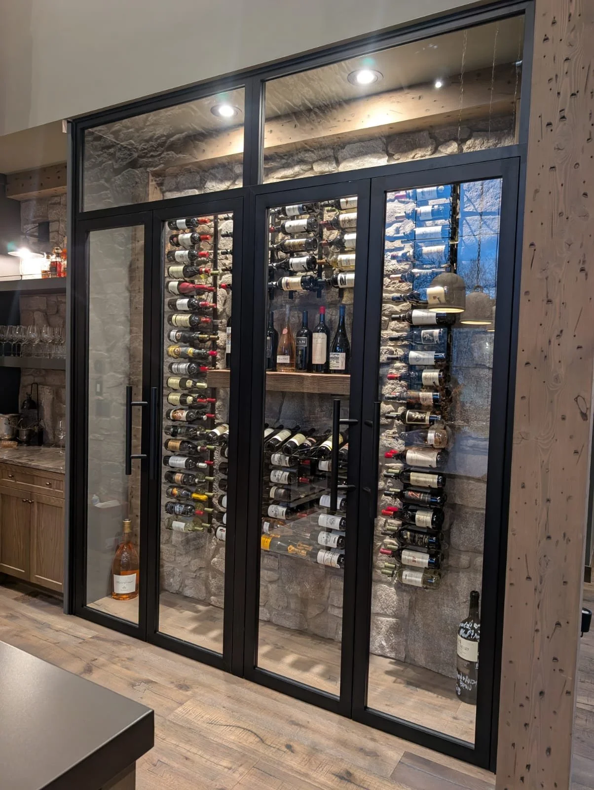 A glass-enclosed wine cellar with walls of stored wine bottles, wooden shelving, and a stone wall background, located inside a modern home or restaurant.
