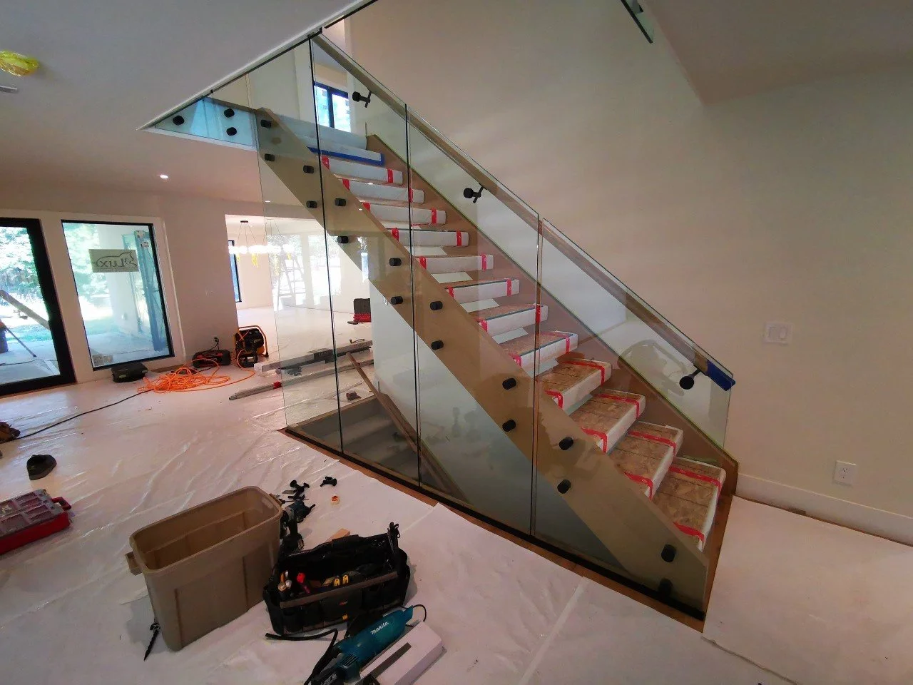 Interior view of a house under construction with a modern glass staircase, construction tools, and supplies scattered on the floor.