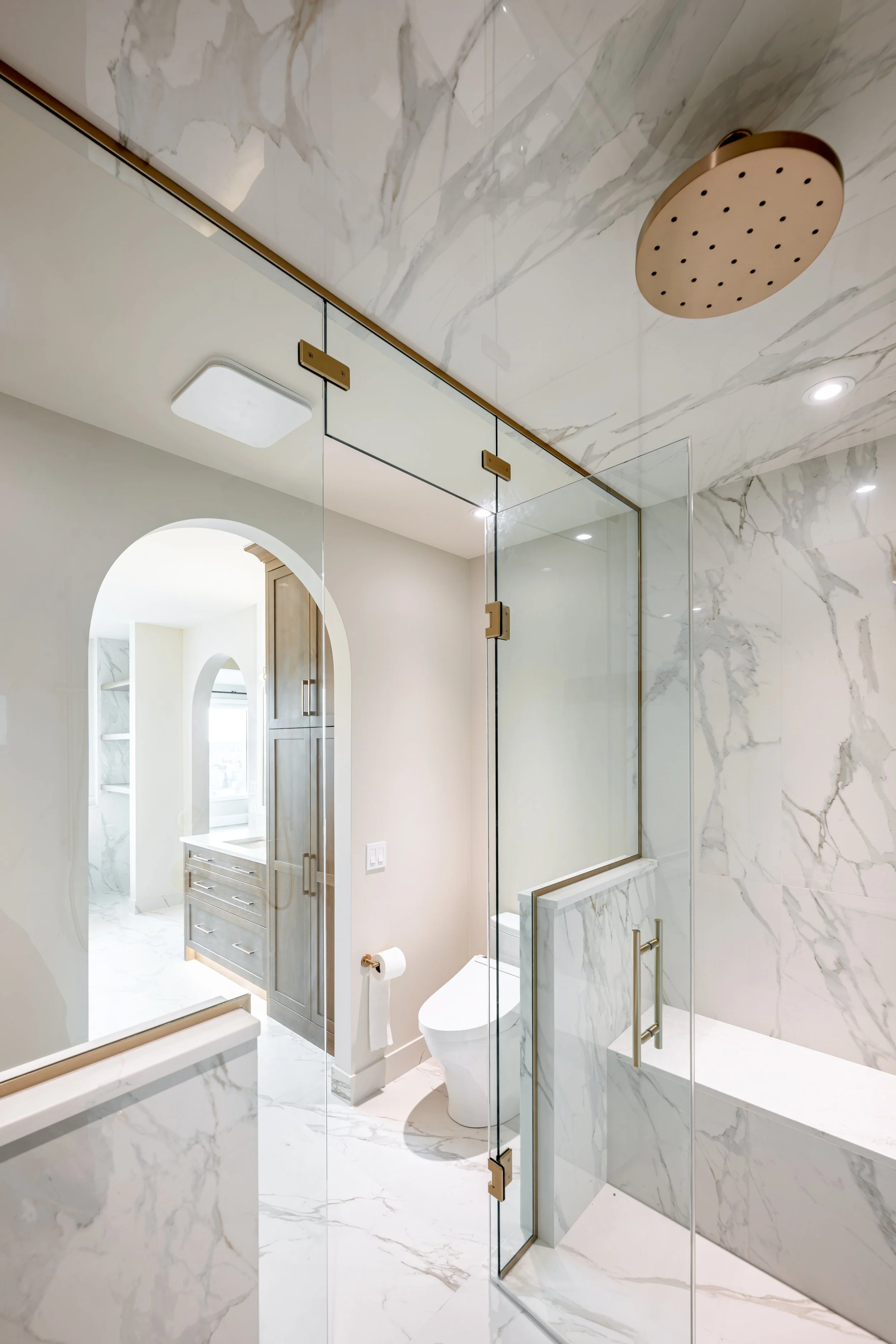 Modern bathroom with marble walls and floors, glass shower enclosure, gold fixtures, and a toilet.