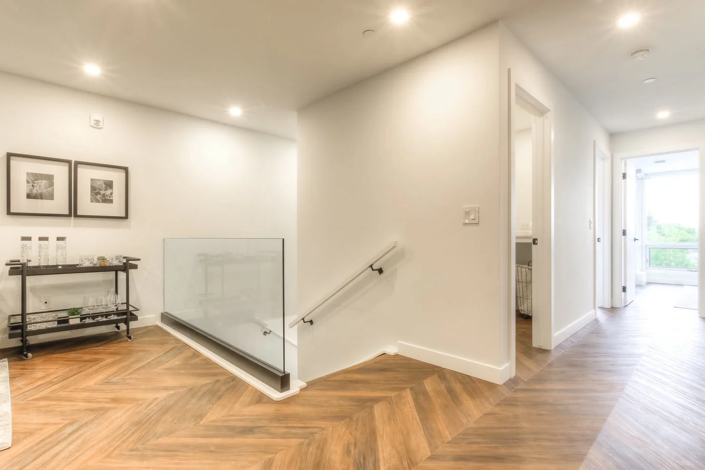 Bright hallway with wooden floor, white walls, and multiple doors. A small bar cart with glassware and a framed black-and-white artwork on the wall. Natural light coming from a large window at the end of the hallway.