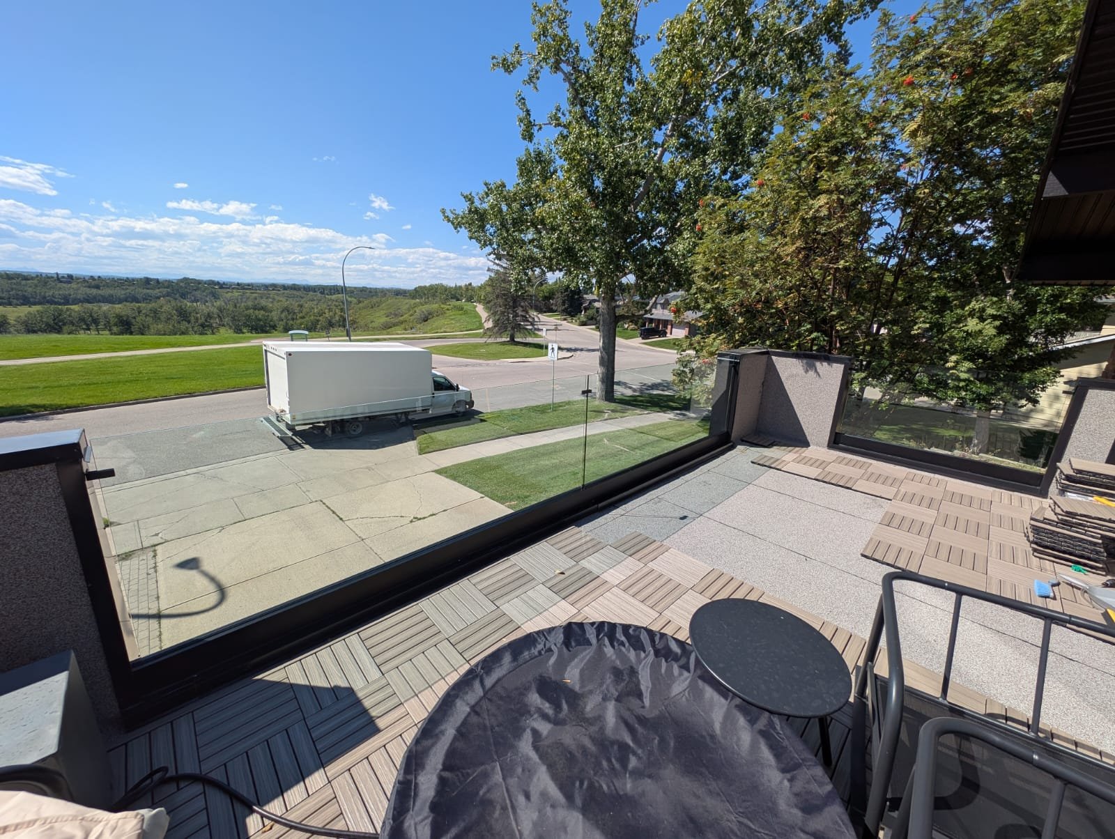 View from a balcony showing a street with a white delivery truck parked, green trees and open fields in the background, and a clear blue sky.