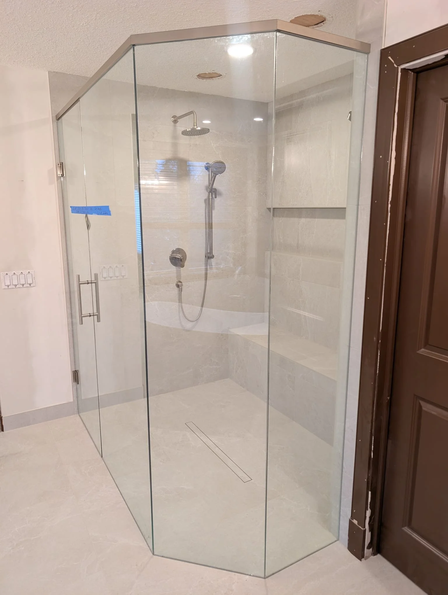 Newly installed clear glass shower enclosure with two walls meeting at a corner, featuring a ceiling-mounted showerhead, a handheld showerhead, and built-in bench, located in a bathroom with beige tile walls and flooring.