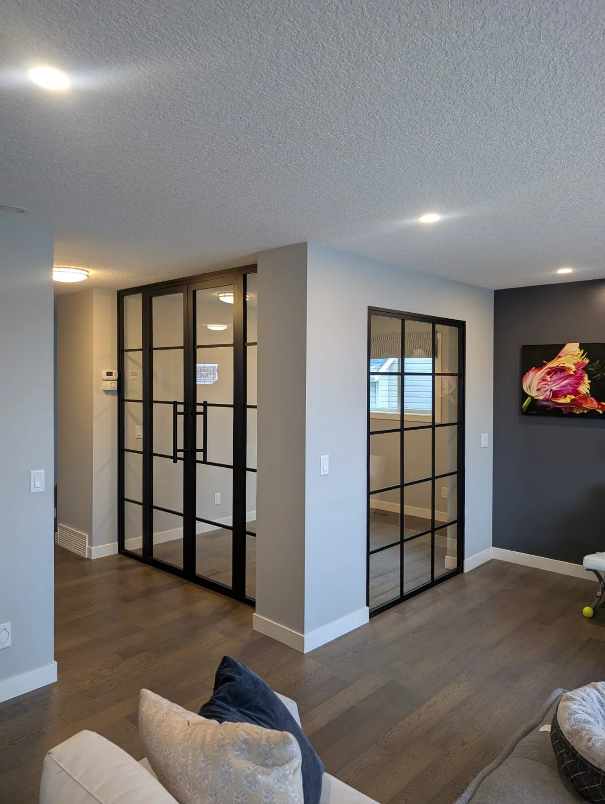 Living room with glass-paneled black framed doors separating the space, hardwood floors, and a dark accent wall with a floral artwork. Interior partition wall.