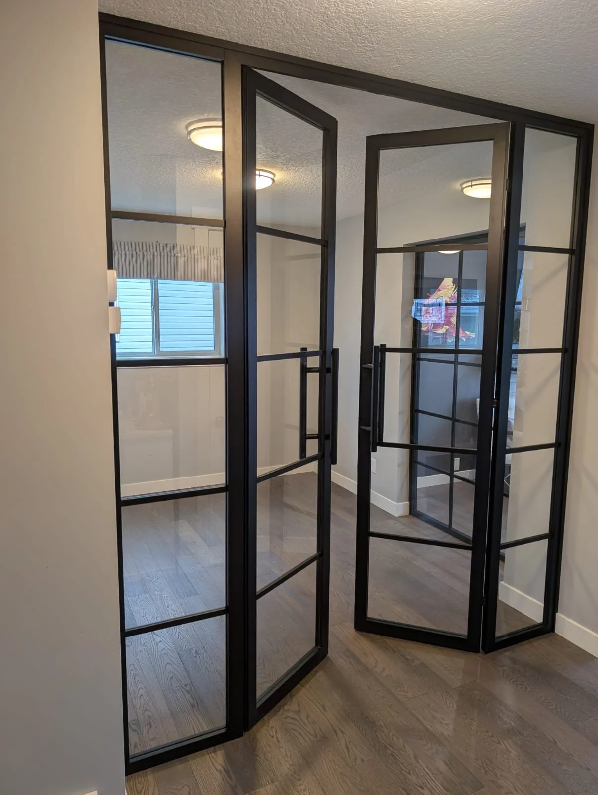 A metal and glass room divider or partition with black frames, set in a room with hardwood floors and white walls, with ceiling lights and a window with blinds in the background.