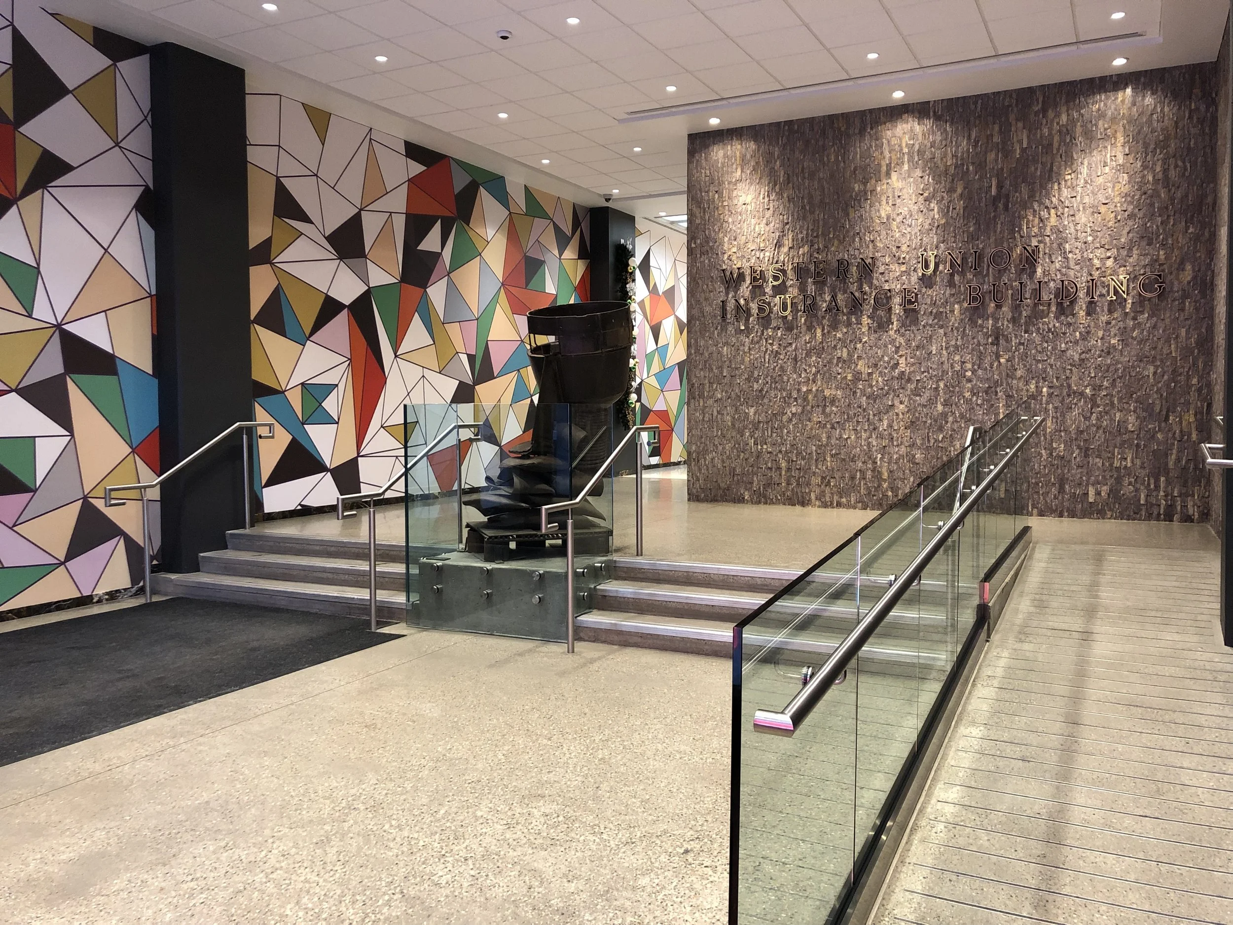 Interior of the Western Union Insurance Building lobby, featuring a colorful geometric mural wall, a stone wall with the building's name, a staircase with glass and metal railings, and a large black sculpture.