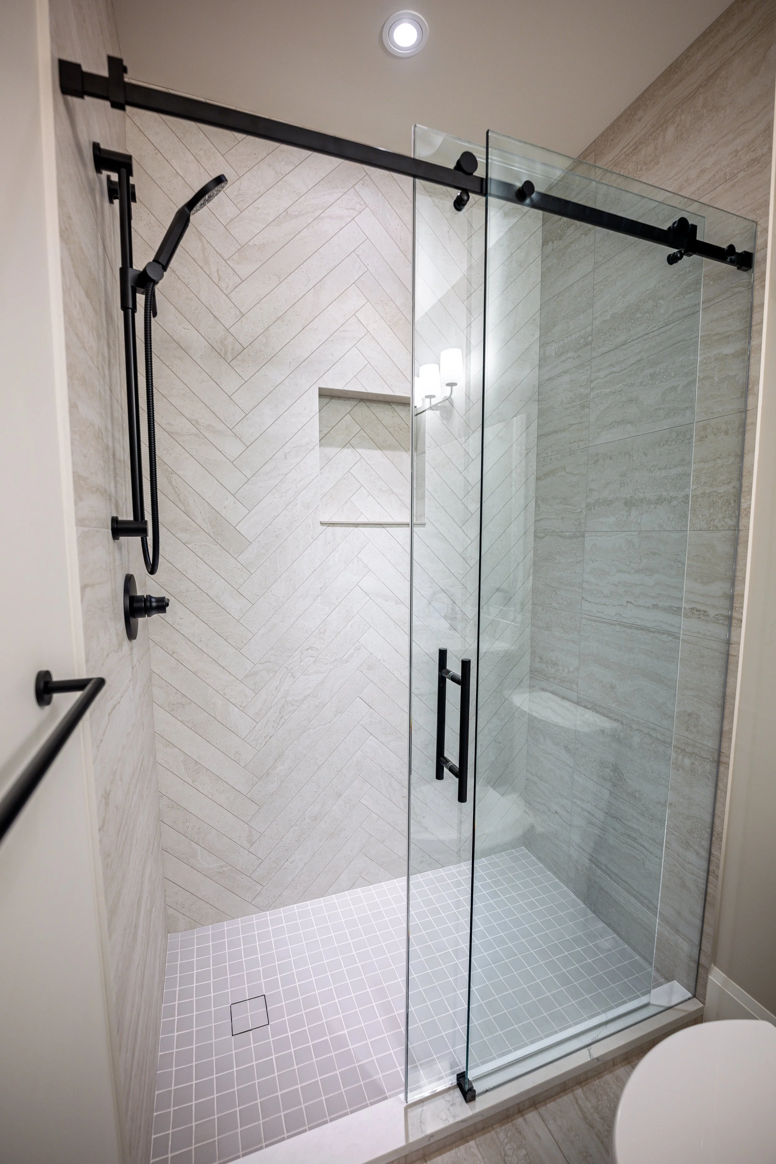 A modern glass shower enclosure with black fixtures, handheld showerhead, and built-in wall niche, inside a bathroom with beige tiled walls and a white tiled shower floor.