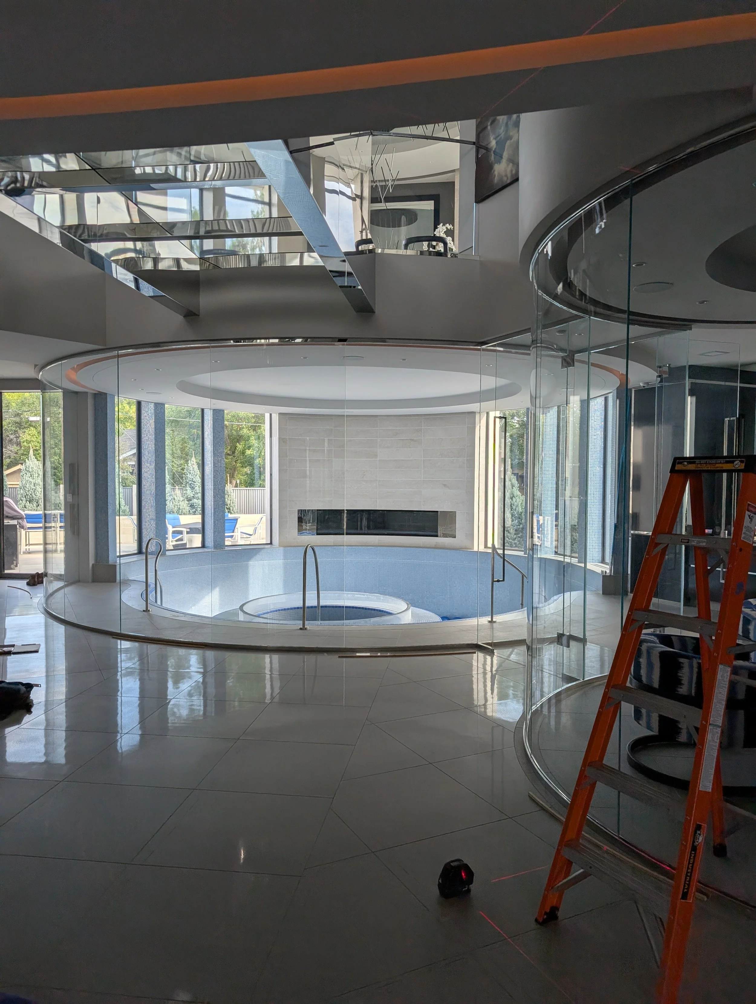 Interior of a modern house with a circular indoor pool, glass walls, and a fireplace, with construction tools including a ladder and laser level on the floor.