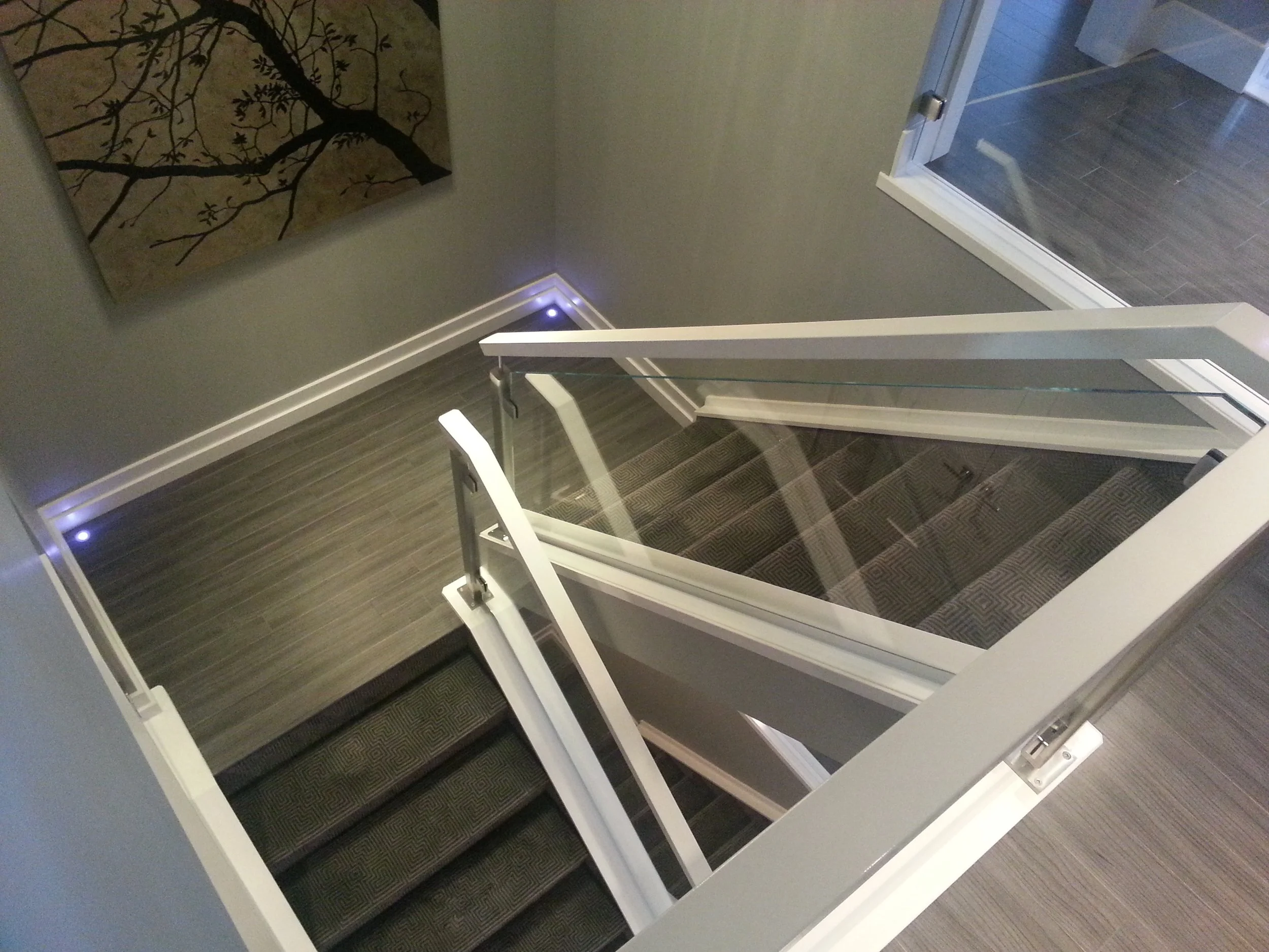 View of stairs and glass railing from above in a modern interior space, with wall art and recessed lighting.
