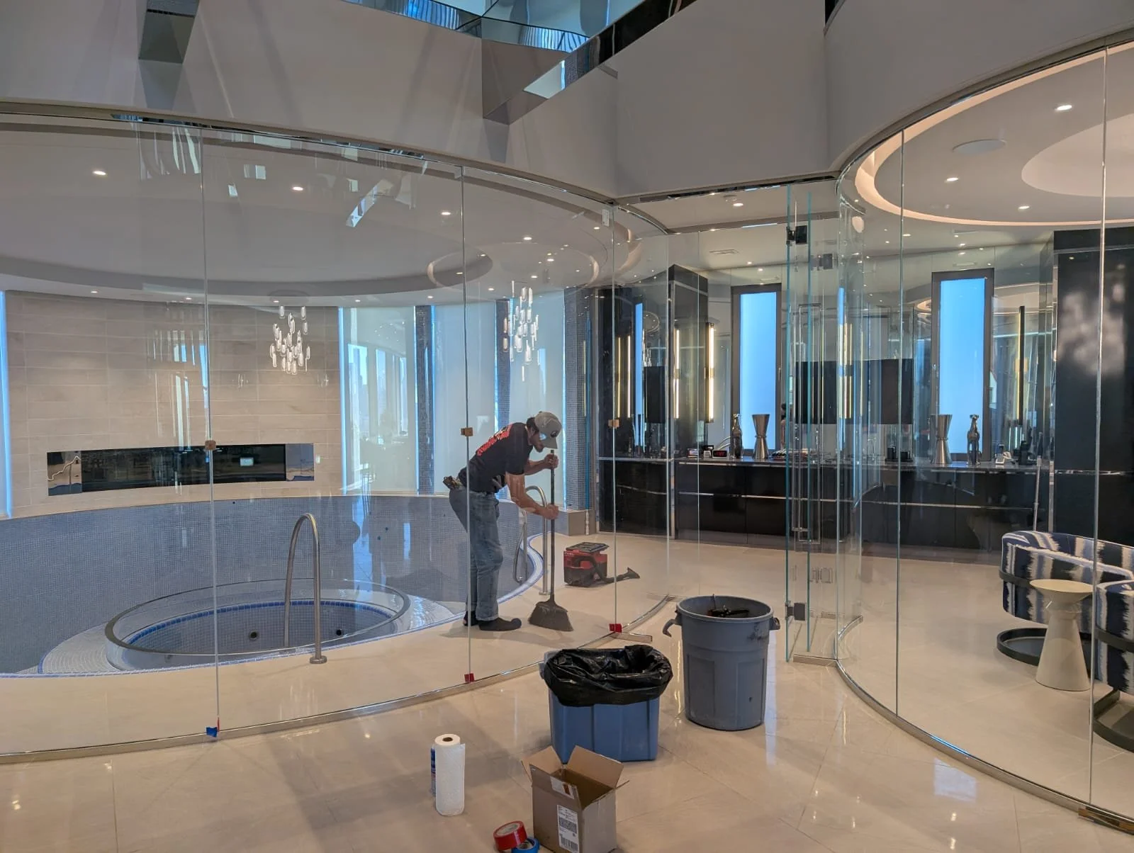 A worker cleaning near an indoor pool area with a glass wall, modern lighting, and decorative vases.