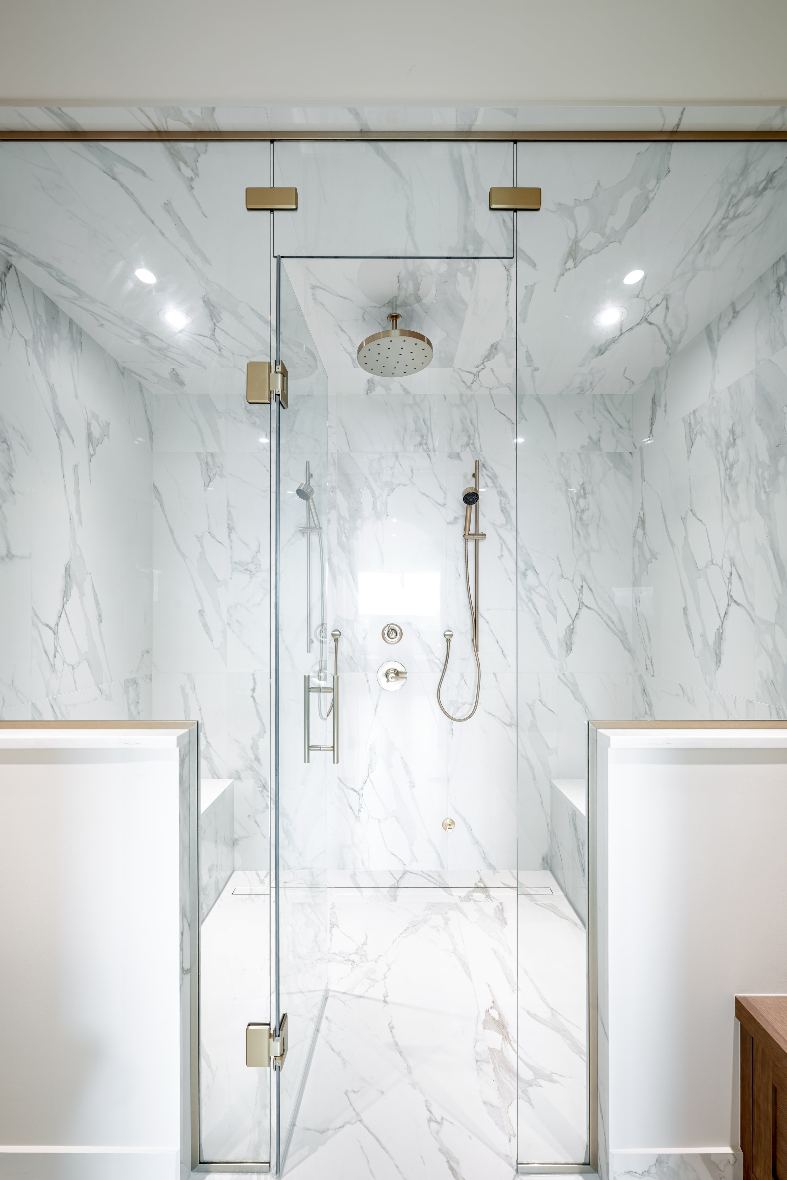 A modern shower enclosure with marble walls and two built-in benches, featuring a rainfall showerhead and handheld shower fixtures.
