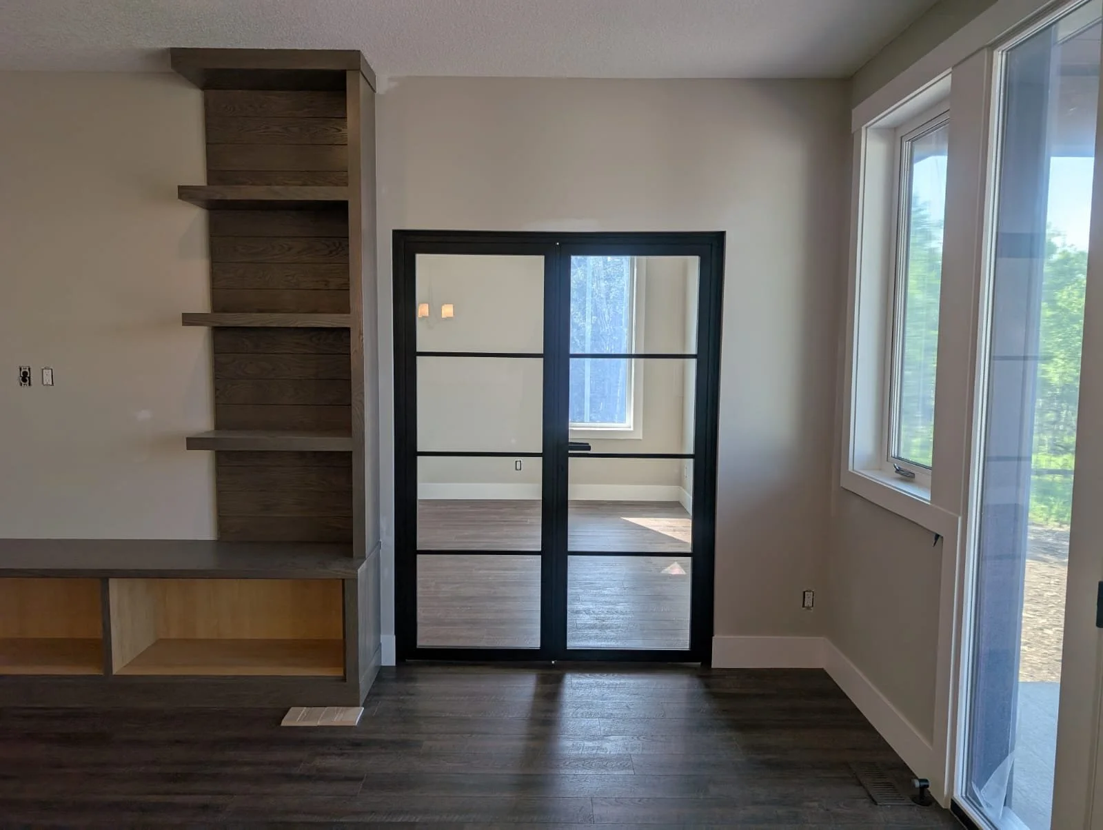 Empty room with dark wood flooring, built-in shelves on the left, aluminum and glass door leading to another room, and a large window on the right showing greenery outside.