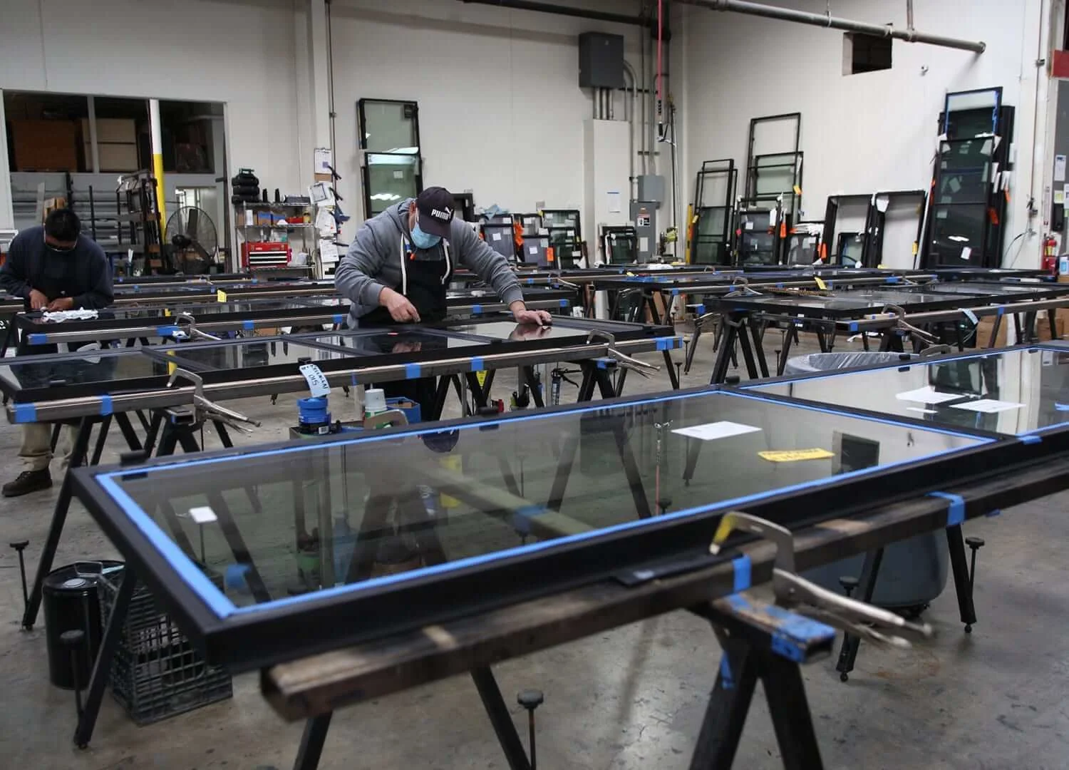 Workers assembling or inspecting large glass panels in a factory setting with multiple workbenches, tools, and equipment in the background.