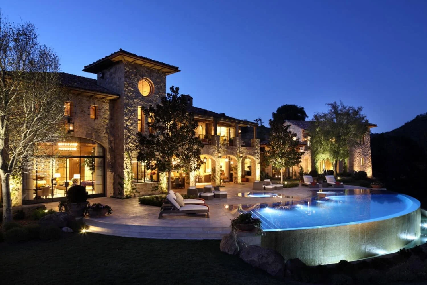 Nighttime view of a luxurious stone mansion with illuminated windows, surrounded by trees, and a lit swimming pool with lounge chairs on the deck.