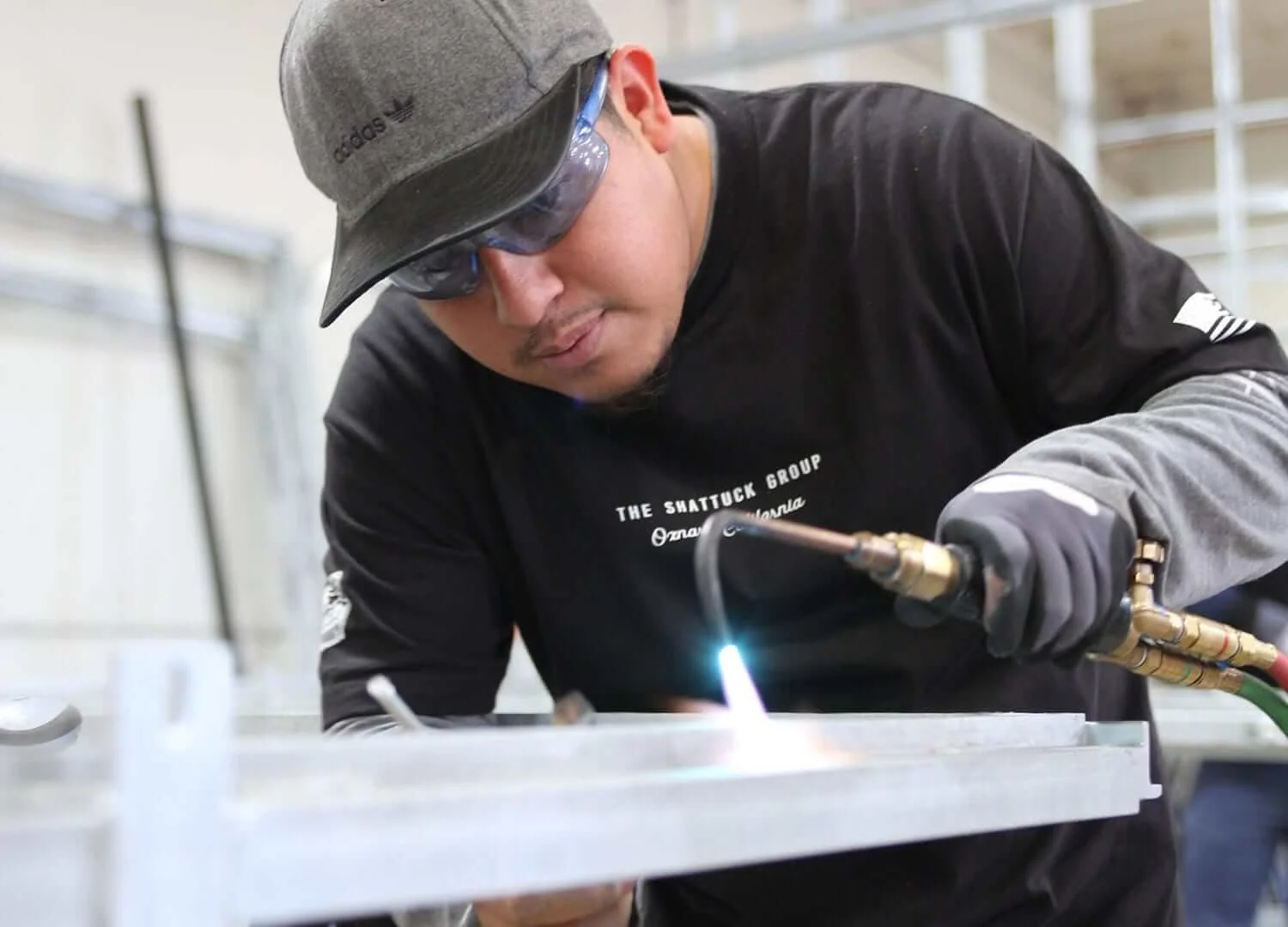 A man welding metal on a workbench, wearing protective gloves, goggles, and a cap indoors.