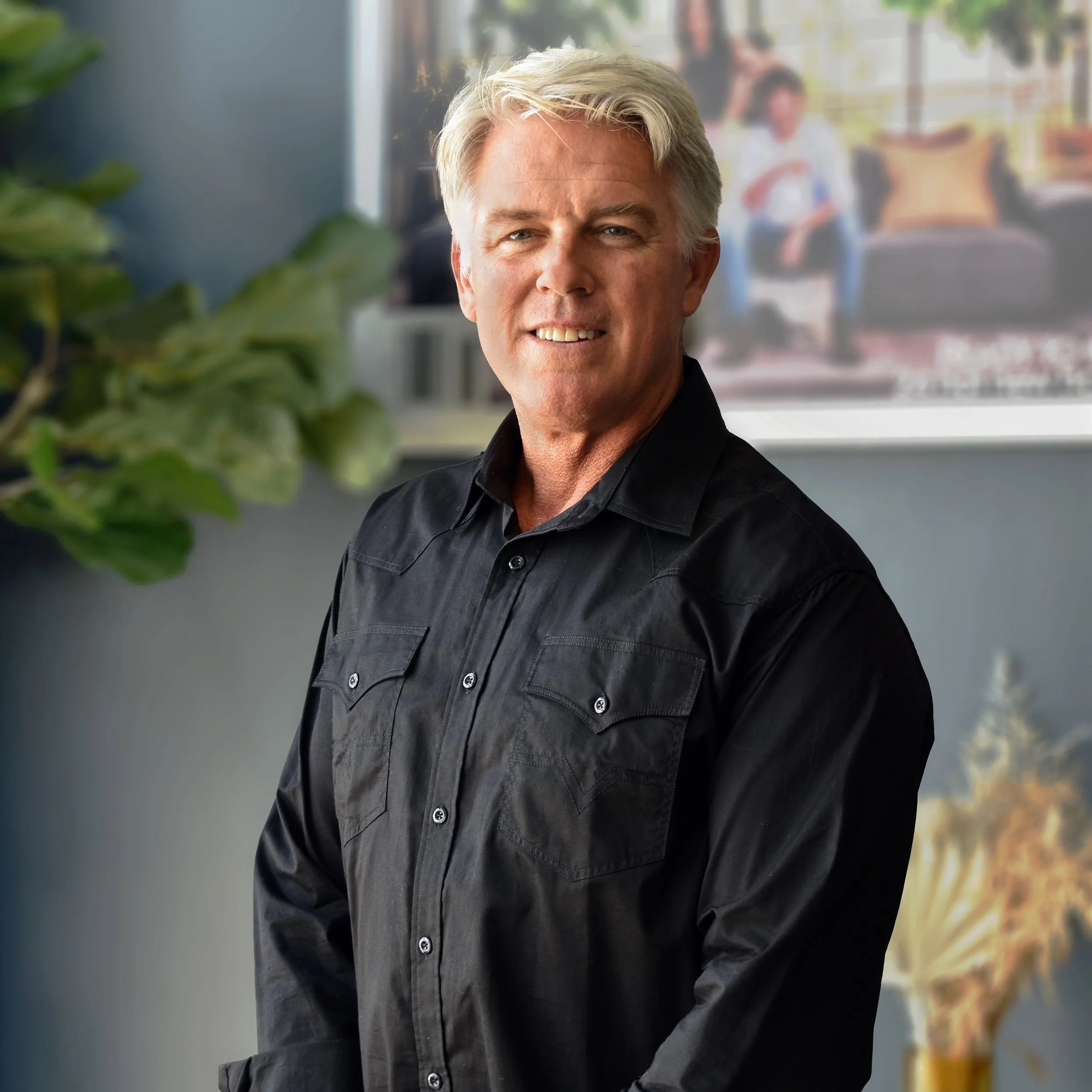 A middle-aged man with gray hair wearing a black button-up shirt, standing indoors with a blurred background showing a woman sitting on a couch and some houseplants.