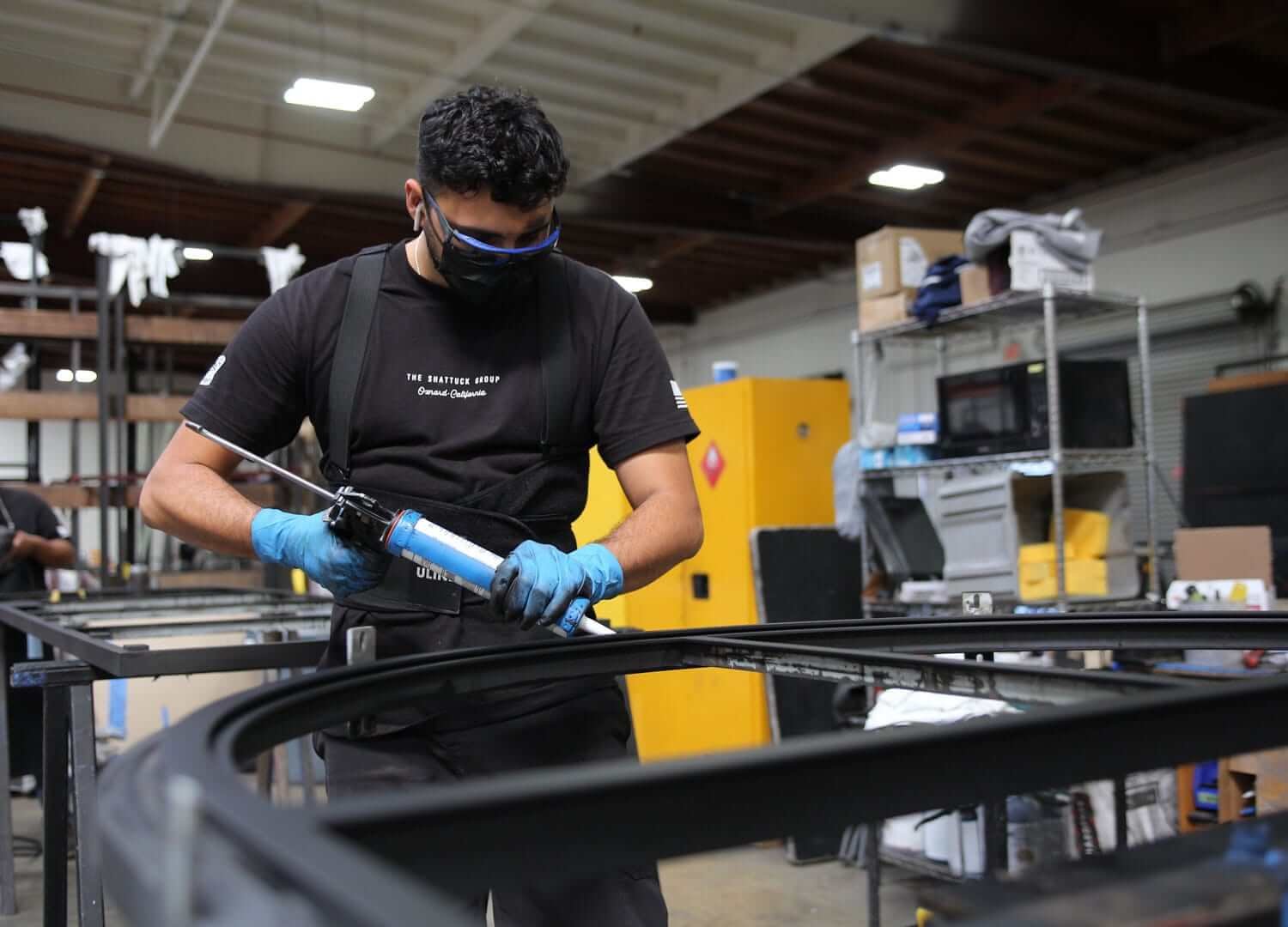 A man wearing a black T-shirt, blue gloves, a face mask, and safety glasses working with a caulking gun on a black metal frame in a workshop.