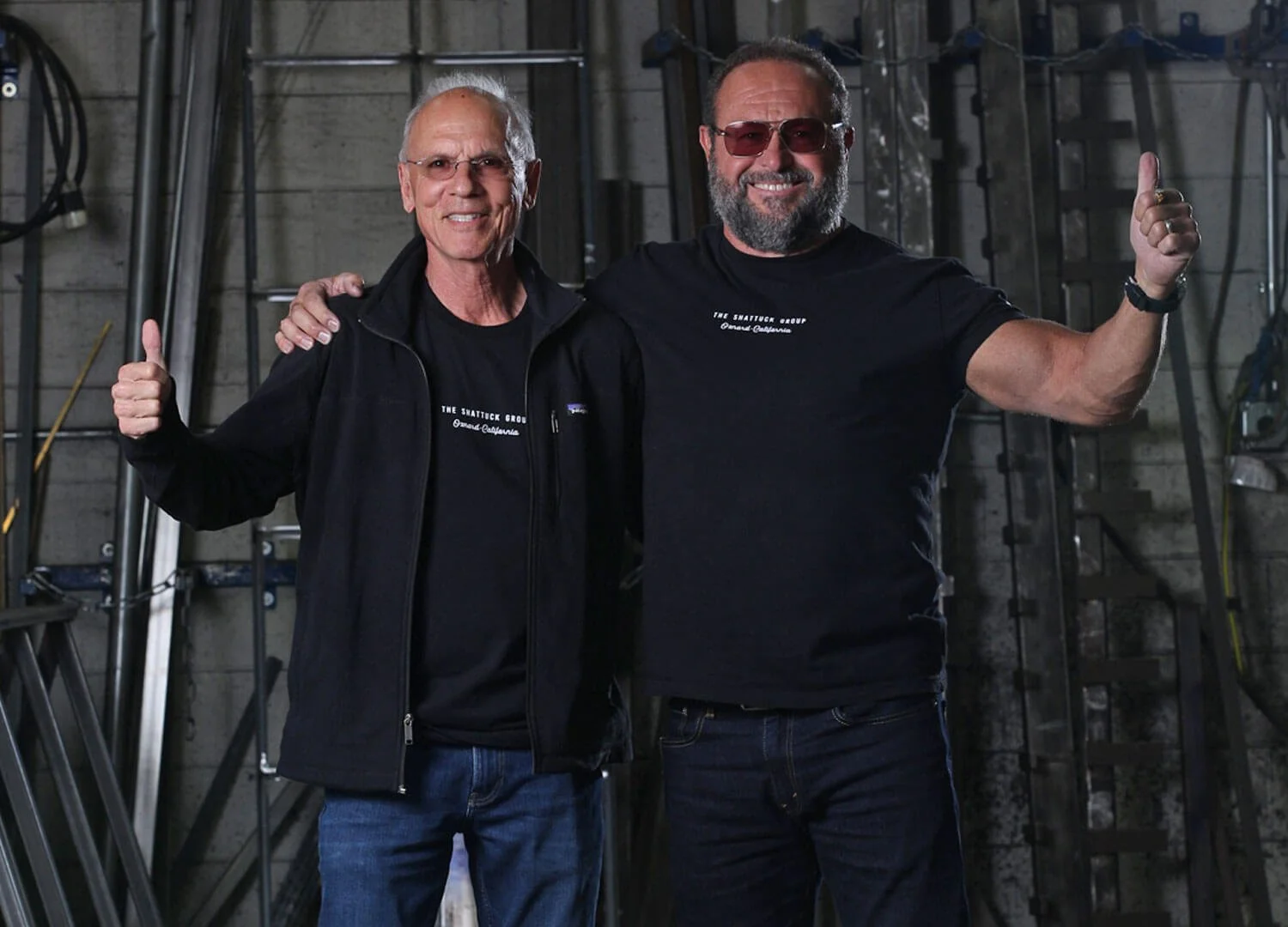 Two men standing close together in an industrial setting, smiling, with one giving a thumbs-up and the other giving a thumbs-up. Both men are wearing black shirts, with one also wearing a black jacket. The background has metal pipes and tools.