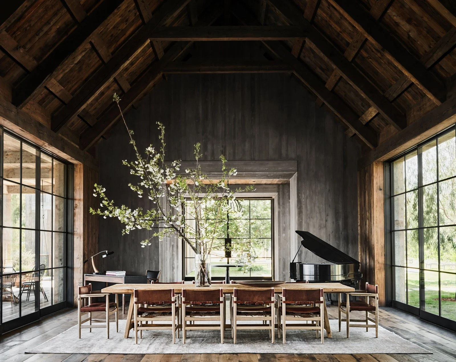 Modern dining room with exposed wooden beams, large windows, a long table with eight chairs, a large vase with flowering branches, a grand piano, and a black desk with a chair.