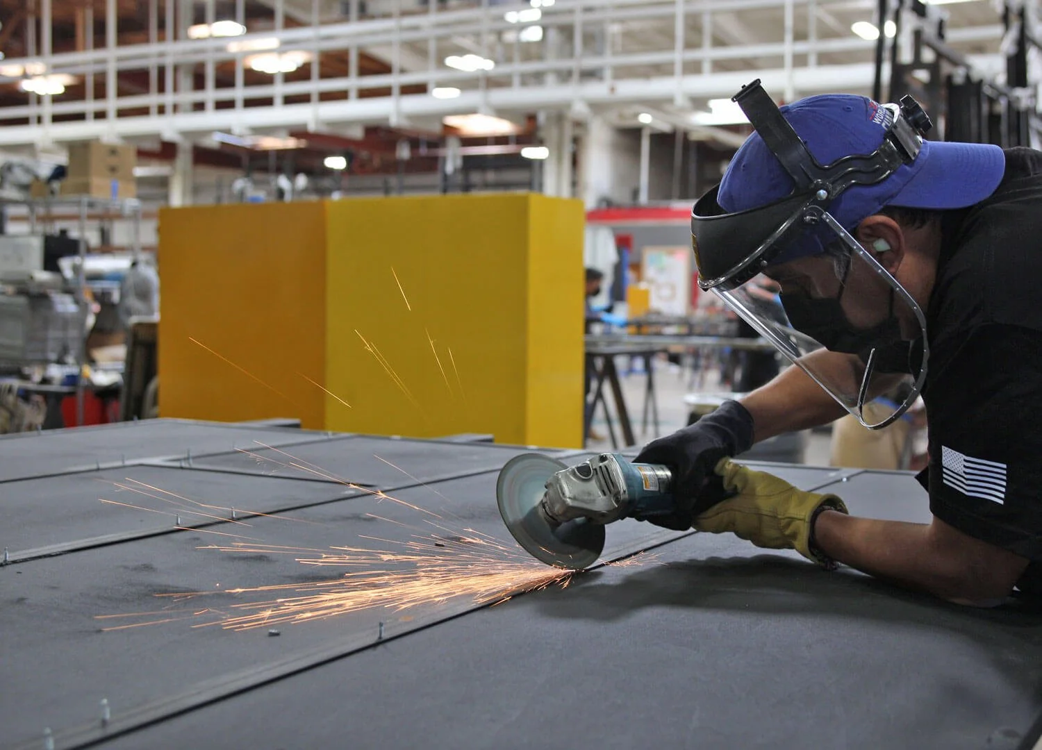 Worker wearing safety gear, including a face shield, grinding metal in an industrial workshop with sparks flying.