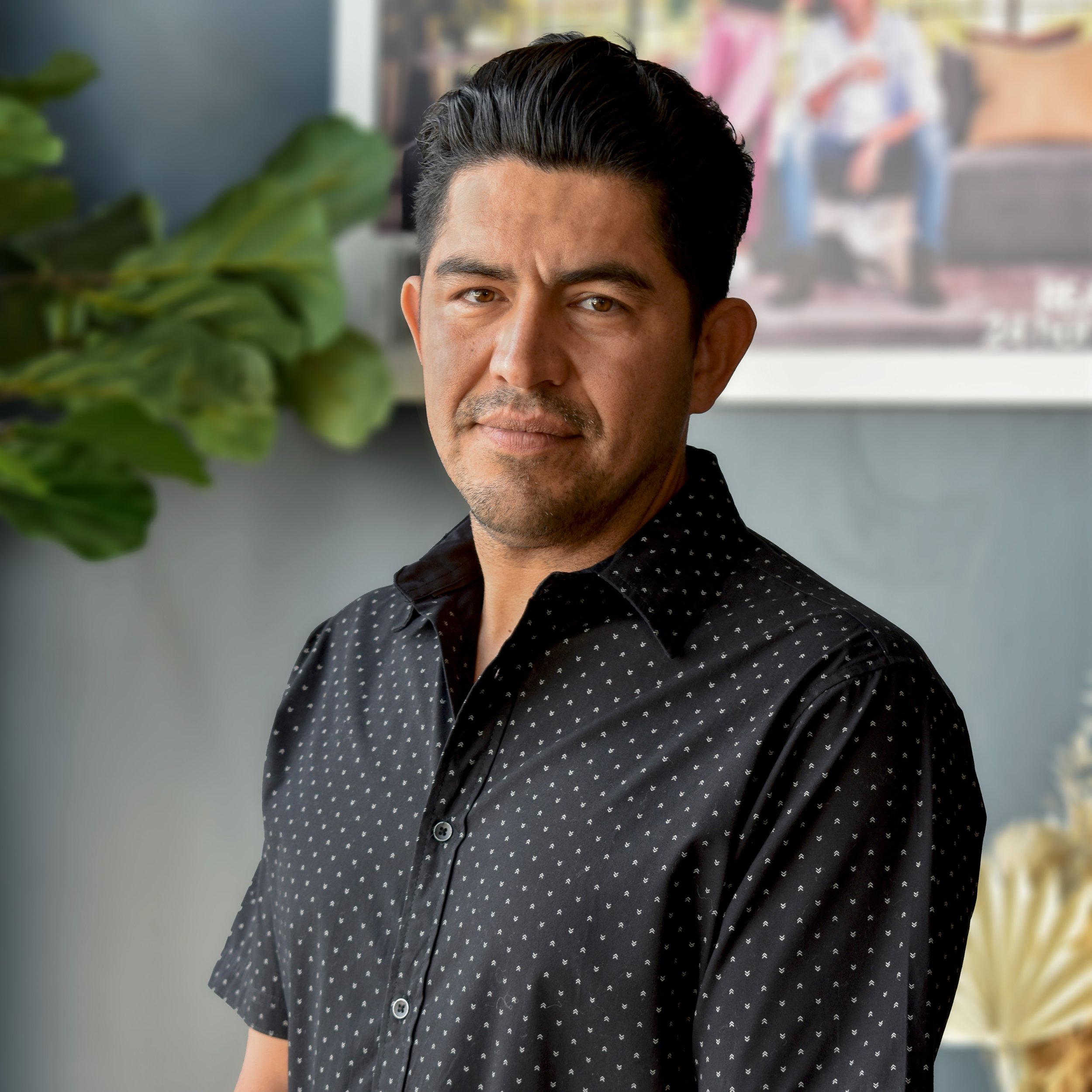 A man with dark hair and a slight beard, wearing a black button-up shirt with small white dots, standing indoors in front of a blurred background with a large green plant and a framed photo or artwork.