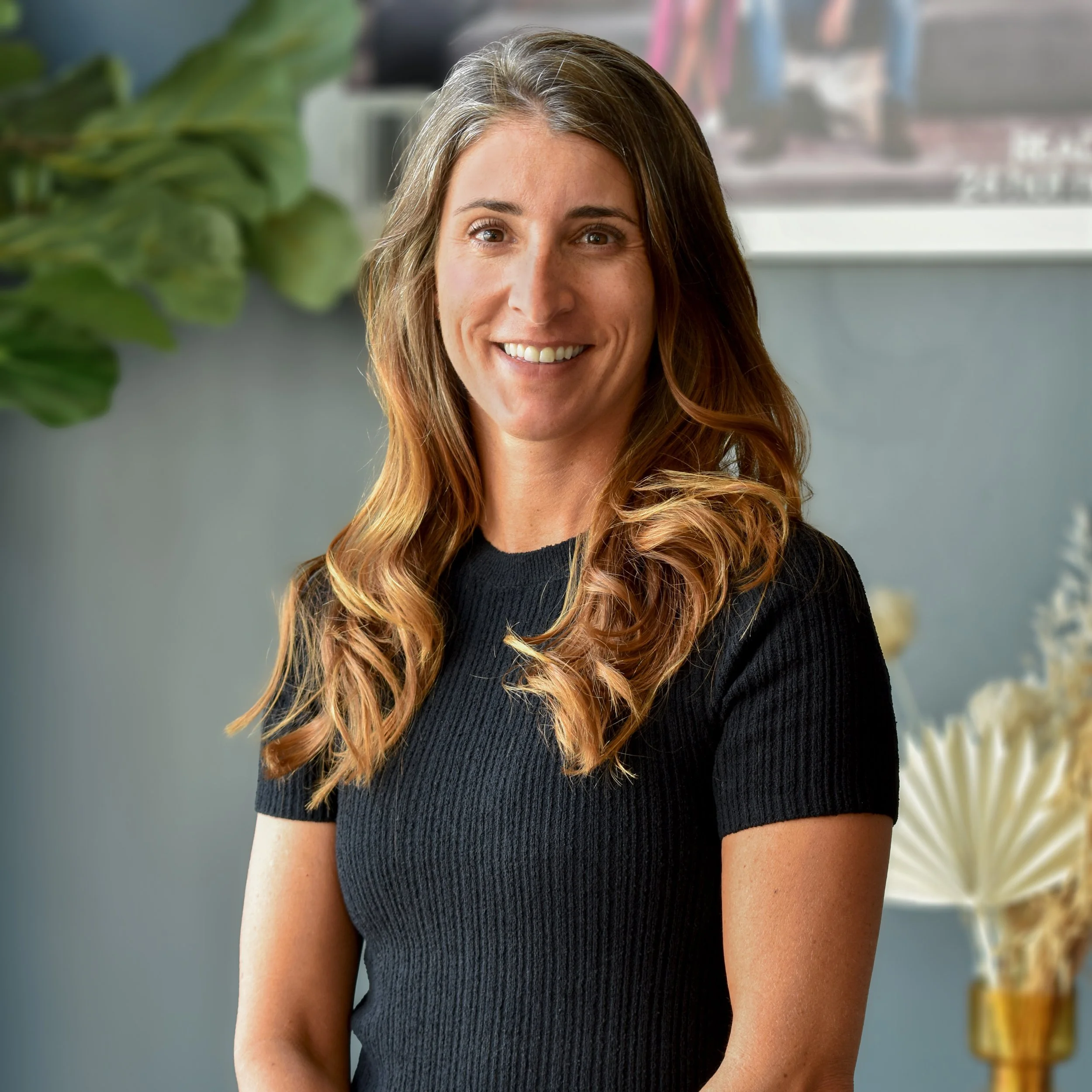 A woman with long, wavy red hair smiling and wearing a black ribbed short-sleeve shirt, standing indoors with a gray wall and green plant in the background.