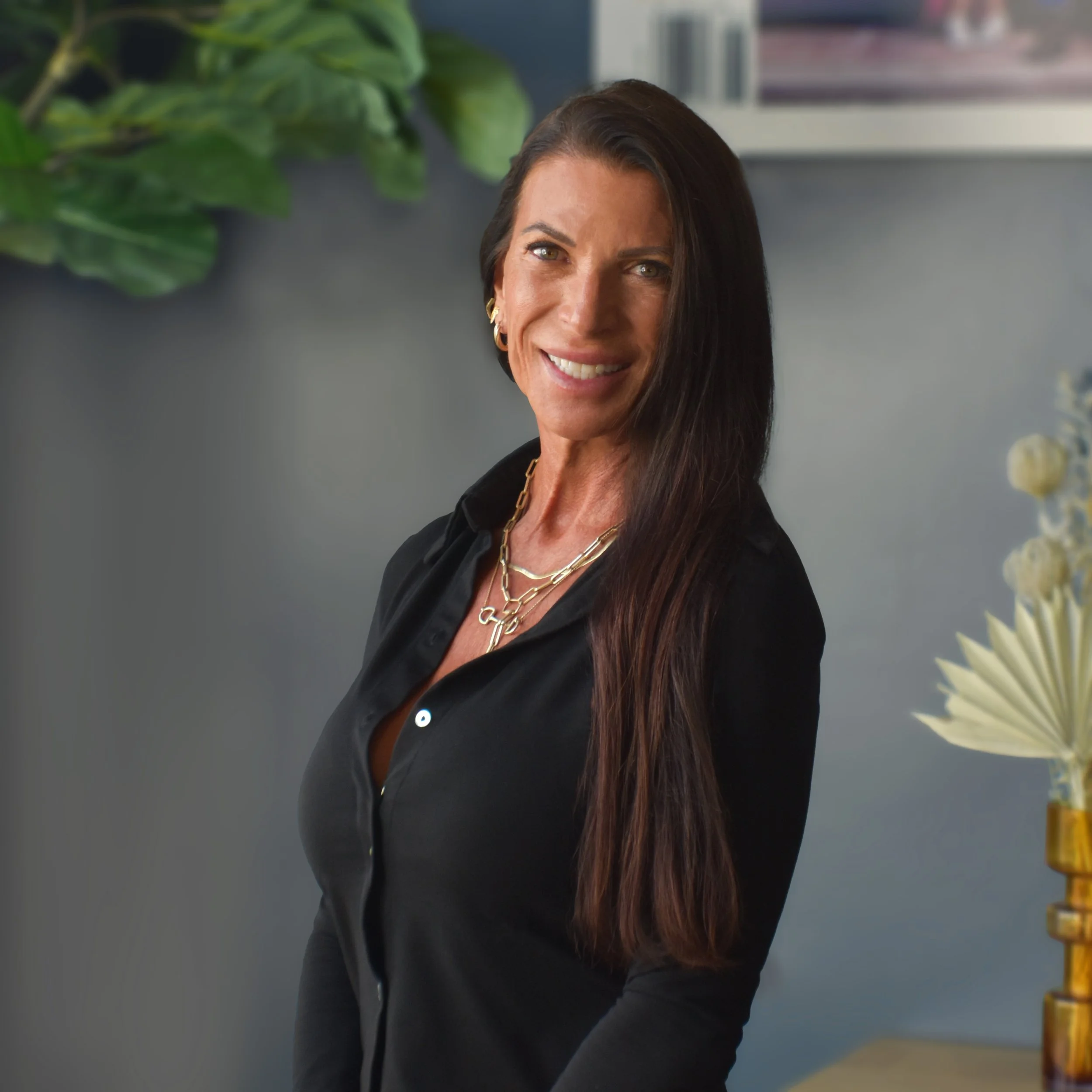 A smiling woman with long dark hair, wearing a black shirt and gold jewelry, standing indoors near a wall with decorative items and plants.