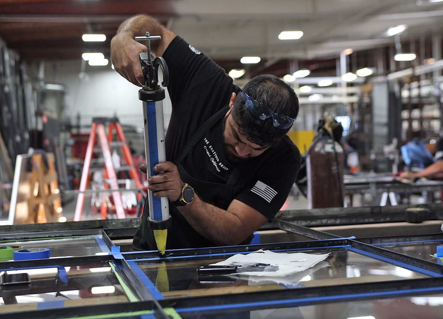 A man working on a large glass panel in a workshop, using a caulking gun. He is wearing safety goggles and a black shirt with an American flag patch on the sleeve.