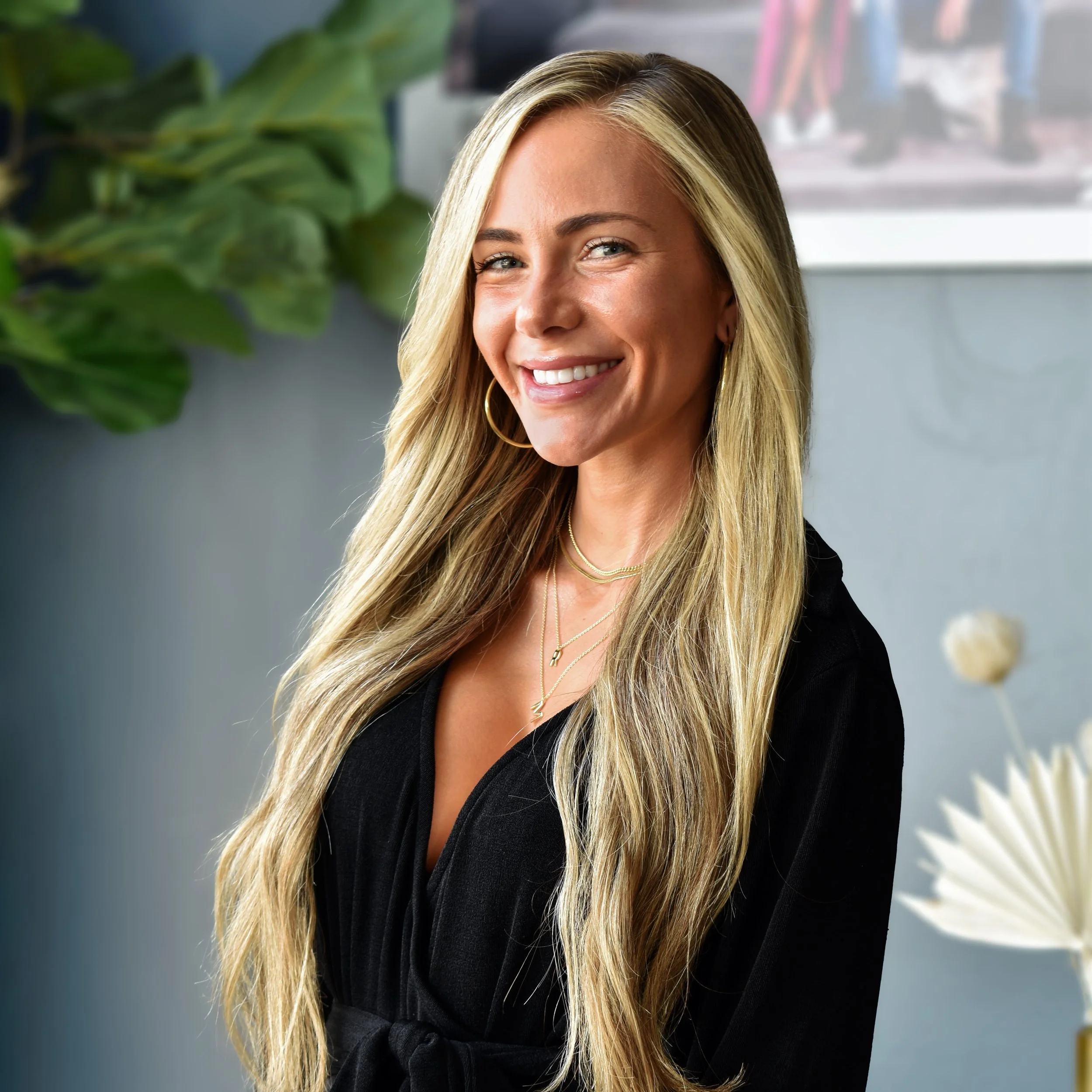 A smiling woman with long blonde hair, wearing gold hoop earrings and layered necklaces, poses indoors against a neutral background with green plants.