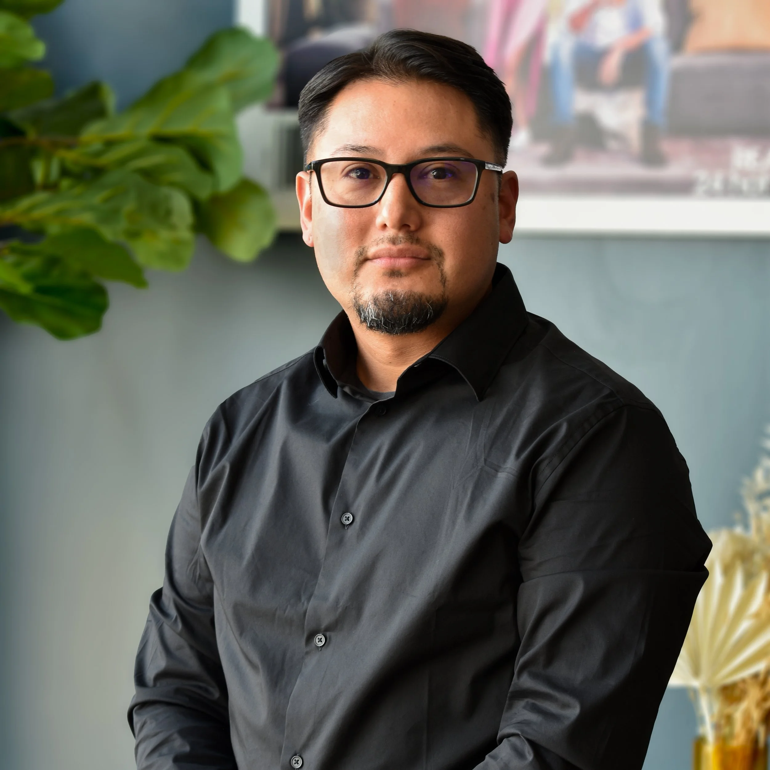 A man wearing glasses and a black shirt, sitting indoors with a large green plant on his left and abstract art in the background.