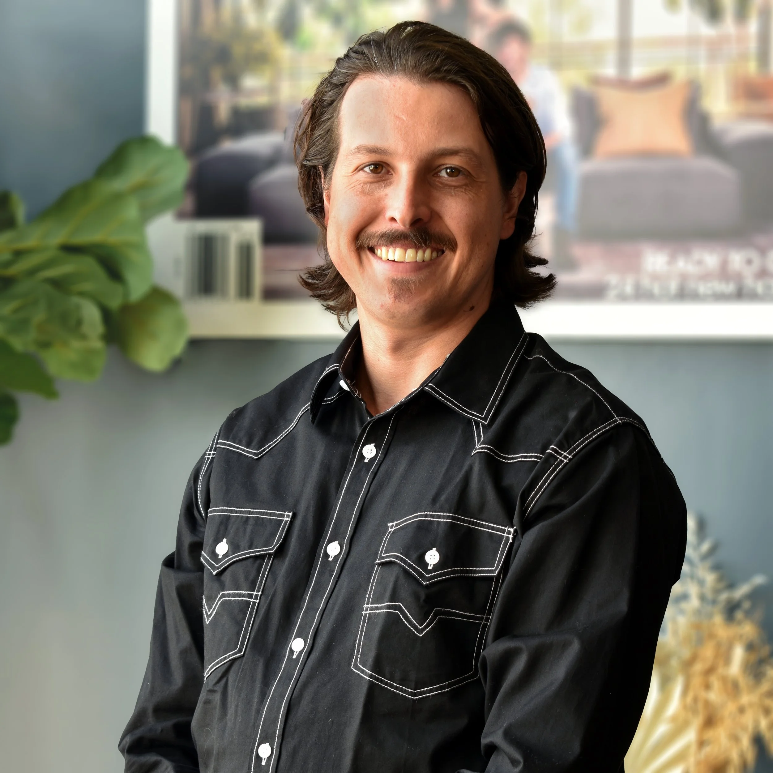 A smiling man with dark hair and a mustache, wearing a black Western-style shirt with white stitching, sitting indoors with a blurred background of a plant and a poster on the wall.