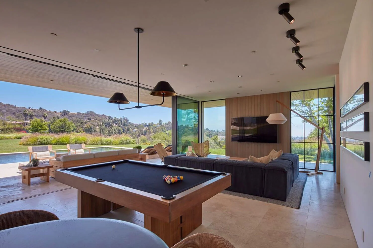 Modern living room with pool table, large windows showing outdoor landscape, sofa, wall-mounted TV, and floor lamp.